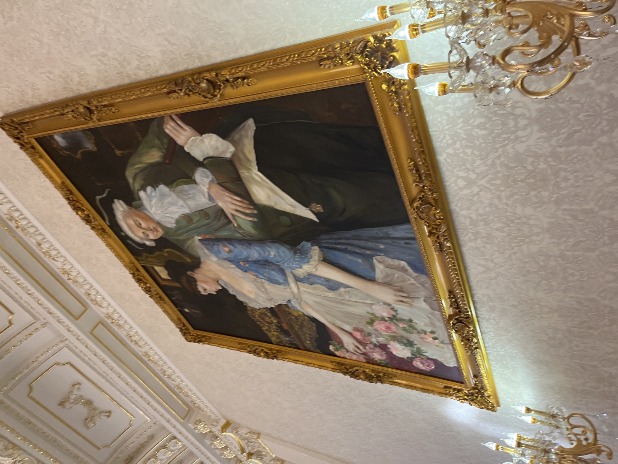 An ornate, gold-framed painting depicting a man and a woman in historical attire hangs on a textured wallpapered wall in a richly decorated room. The man, on the right, wears a green jacket with ruffled white cuffs and collar, and a powdered wig. The woman, on the left, is in a blue dress with a white apron, and she is reaching towards a bouquet of pink and white flowers. The room features elaborate white and gold decorative molding on the ceiling. To the left and right of the painting are crystal chandeliers with multiple candle-shaped lights. The scene is illuminated by the lights of the chandeliers and possibly ambient light from an unseen source. The perspective is from a low angle, looking up at the painting and the ceiling. This painting is located in the Dubivtsi, Ukraine area.