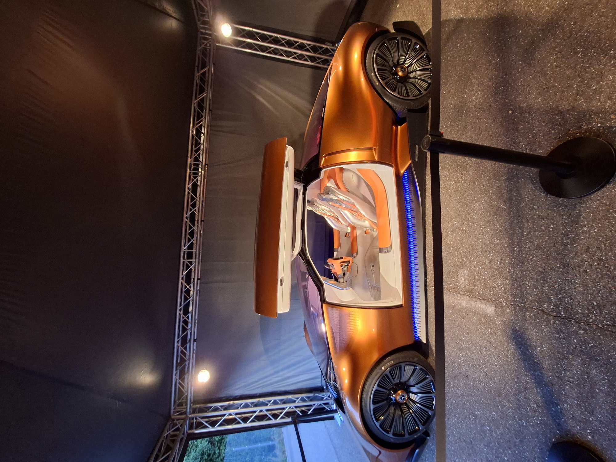 The image shows a futuristic concept car, likely the Mercedes-Benz Vision AVTR, displayed outdoors under a tent structure. The car is a vibrant orange color with a sleek, aerodynamic design. It features gull-wing doors that are open, revealing a minimalist interior with white and orange seats and a central control panel. The car has distinctive wheels and a large rear spoiler that opens upwards. Blue LED lighting illuminates the lower sides of the car. The car is situated on a dark asphalt surface, and a black stanchion with a red ribbon is visible in the foreground, suggesting a cordoned-off exhibit. In the background, a dark fabric backdrop is visible, supported by metal truss structures with spotlights. To the left, a glimpse of greenery and a fence suggests an outdoor setting, potentially near a road or pathway. The overall impression is of a technology showcase or automotive exhibition. The provided information indicates the location is Rastatt, Germany.