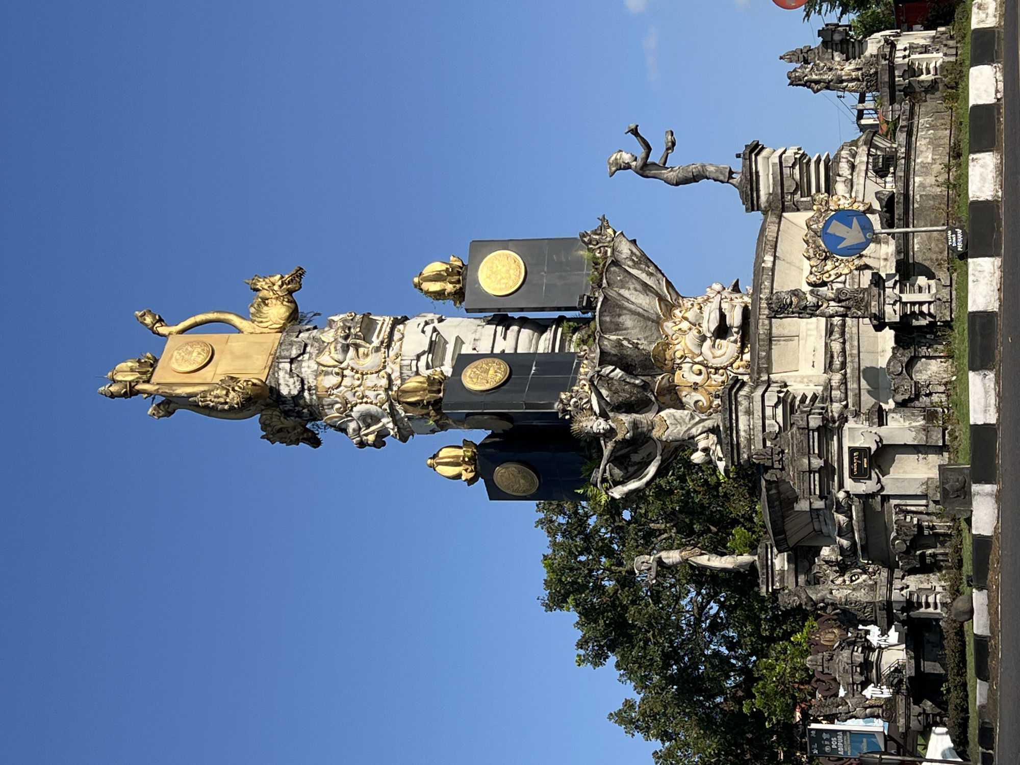 The image shows the Catur Muka Monument in Tabanan, Bali, Indonesia, under a clear blue sky. This monument is a circular roundabout featuring a large, elaborate sculpture at its center. The sculpture is adorned with gold accents and depicts several figures. At the very top, there's a golden serpent-like creature with a disc. Below this, several large black pillars with golden discs are stacked, topped with golden crowns.
The base of the sculpture features multiple stone statues. On the left, a female figure with outstretched arms is depicted, adorned with intricate clothing and headwear. On the right, a male figure stands holding a bowl and a tool, also in traditional attire. Various smaller stone carvings and architectural elements form the foundation of the monument, including what appear to be mythological creatures or deities.
The roundabout itself has a paved road surface with white and black striped markings along the edge. A blue circular sign with a white arrow pointing downwards is visible in front of the monument, indicating a traffic direction. Green grass and shrubs surround the base of the monument. In the background, lush green trees partially obscure some of the surrounding environment. To the far left, a portion of a white sign with blue text is visible. The overall scene captures a prominent landmark in Tabanan, characterized by its rich cultural and artistic details.