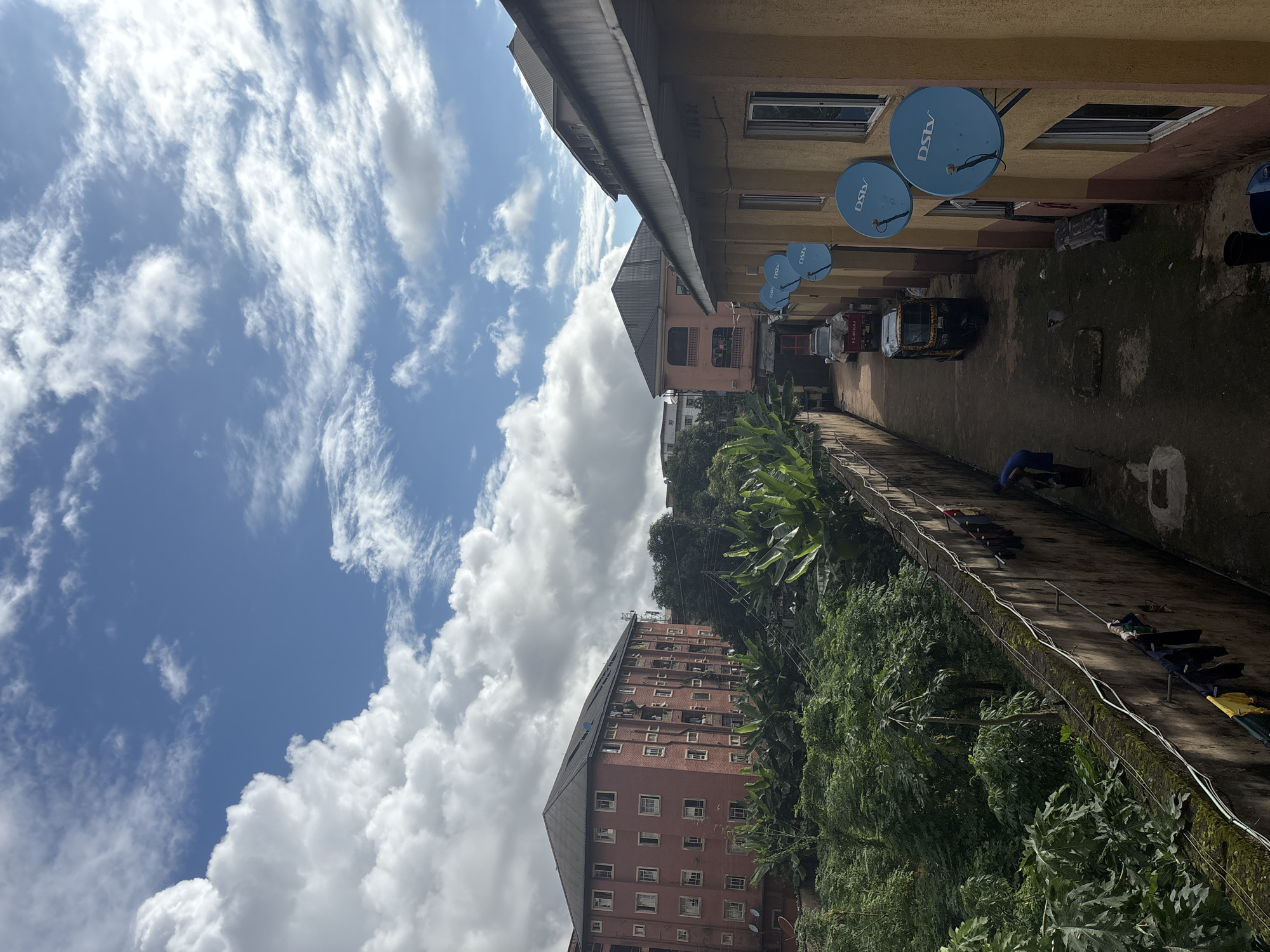 The image depicts an alleyway in the city of Onitsha, Nigeria, with buildings on either side and a view of the sky above. On the right, a two-story building with yellow and brown painted walls features multiple DStv satellite dishes mounted on the exterior. The building has several windows with white frames. In the alley, a yellow auto-rickshaw, also known as a "keke," is parked, and another vehicle is partially visible behind it. A person wearing a blue garment is standing near the auto-rickshaw. On the left side of the alley, a tall, weathered concrete wall runs along the length of the scene, with clothes hanging on a line attached to it. Beyond the wall, lush green vegetation and trees are visible, and in the distance, a multi-story pink apartment building stands. The sky is a vibrant blue, dotted with large, fluffy white clouds. The scene appears to be a residential or mixed-use area, common in urban environments like Onitsha.