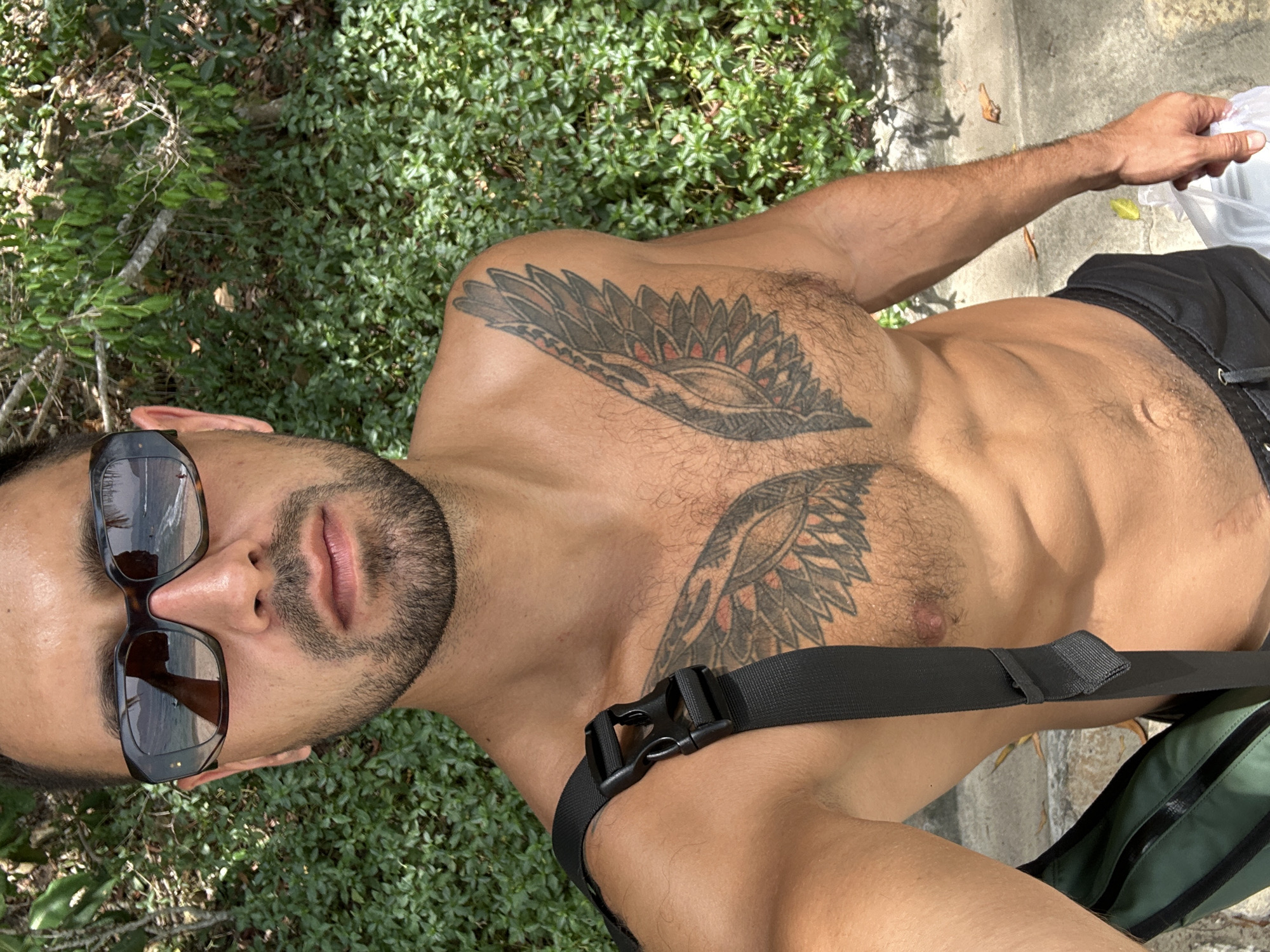 A topless man with a beard, wearing sunglasses and black swim trunks, stands outdoors in Nha Trang, Vietnam. He has a tattoo of wings on his chest and is carrying a green bag over his shoulder. The background is filled with lush green foliage. The scene appears to be a sunny day, likely near a beach, given the reflection of the sea and sand in his sunglasses.