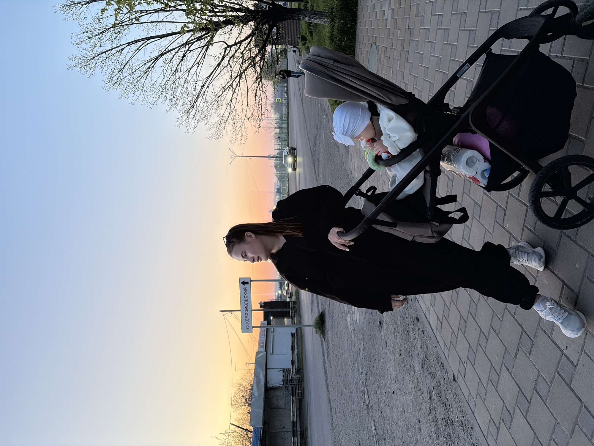 The image captures a peaceful twilight scene in Novopokrovskaya, Russia, featuring a woman pushing a baby in a stroller along a paved sidewalk. The woman, dressed in a black jacket and pants with white sneakers, walks towards the left of the frame, holding the stroller's handle. Her dark hair is pulled back, and her gaze is directed forward. Inside the dark-colored stroller, a baby or small child, wearing a white bonnet and light clothing, is visible, engaged with a colorful toy. A bottle of water and possibly other items are stored in the basket beneath the stroller.
The time of day is sunset or twilight, evidenced by the gradient sky transitioning from warm orange and yellow hues near the horizon to a lighter blue overhead. Bare tree branches with new buds are silhouetted against the sky in the upper left. In the background, an asphalt road runs parallel to the sidewalk, with a car visible in the distance, its headlights illuminated. Several buildings, utility poles, and wires are part of the urban landscape.
A prominent white sign with a directional arrow is visible behind the woman, displaying Cyrillic text. The upper line reads "пер. КОМСОМОЛЬСКИЙ" (per. Komsomolsky), indicating "Komsomolsky Lane". Below it, a partial sign reads "МИР ЦВЕТОВ" (Mir Tsvetov), meaning "World of Flowers," suggesting a flower shop nearby. The setting appears calm and serene during the fading daylight.