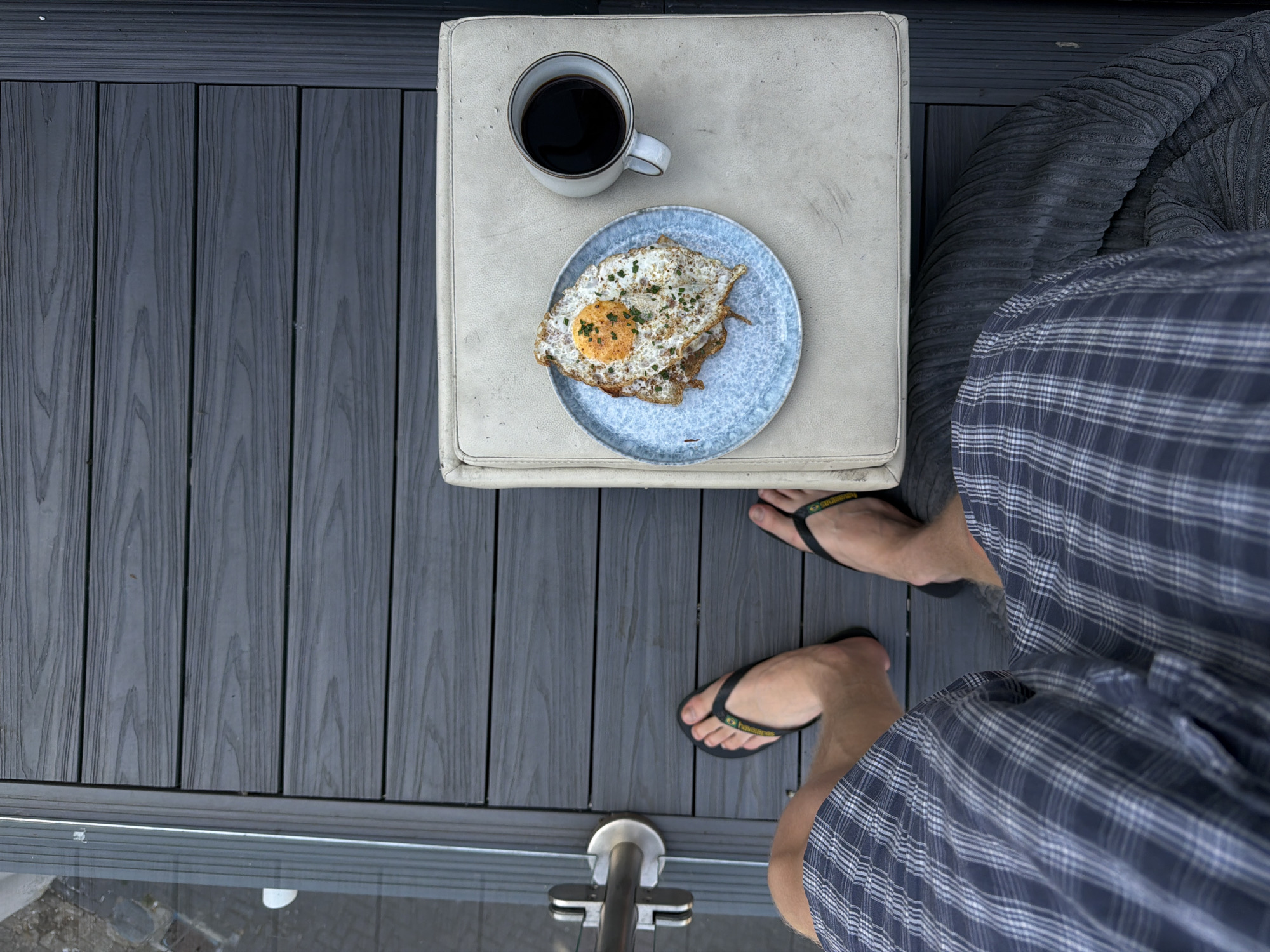A high-angle shot shows a person sitting on a padded outdoor seat on a dark wood deck. The person is wearing plaid shorts and black flip-flops. In front of them, on a small light-colored table, is a plate with two fried eggs and a mug of black coffee. To the left, a glass railing is visible. The scene appears to be on a balcony or patio.