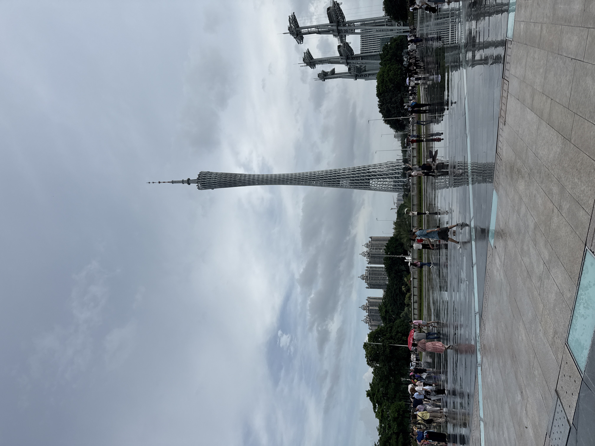 This image depicts the Canton Tower in Guangzhou, China, located in the Tianhe District. The tower, a prominent landmark, stands tall and slender against a cloudy sky. In the foreground, a large, wet, paved plaza reflects the sky and the surrounding structures. Numerous people are scattered across the plaza, some holding umbrellas, indicating recent or ongoing rainfall. Several individuals are walking, while others are standing or gathered in groups. Behind the plaza and the tower, trees and modern buildings form the urban landscape. To the right of the Canton Tower, a set of tall, industrial-looking structures with multiple levels and suspended platforms are visible. The overall atmosphere suggests a public gathering space after a period of rain.
