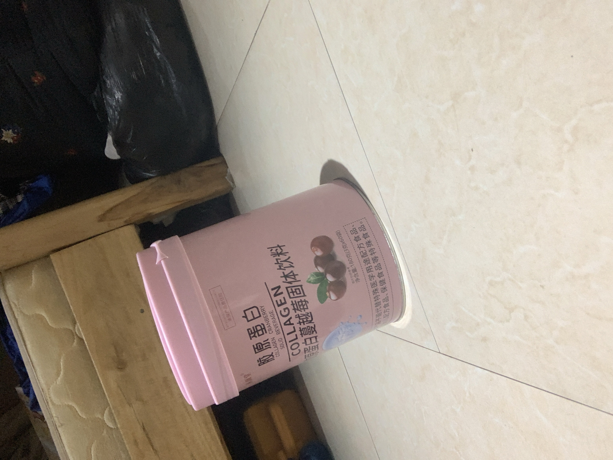 A pink cylindrical container of "COLLAGEN CRANBERRY SOLID BEVERAGE" is placed on a light-colored tiled floor. The container has Chinese characters and English text on its label, along with an illustration of cranberries. The scene appears to be indoors, with a wooden bed frame and bedding visible in the background, and a black plastic bag to the right. The lighting is dim. There is no indication of the city of Abuja, Nigeria, or any specific geographical markers.