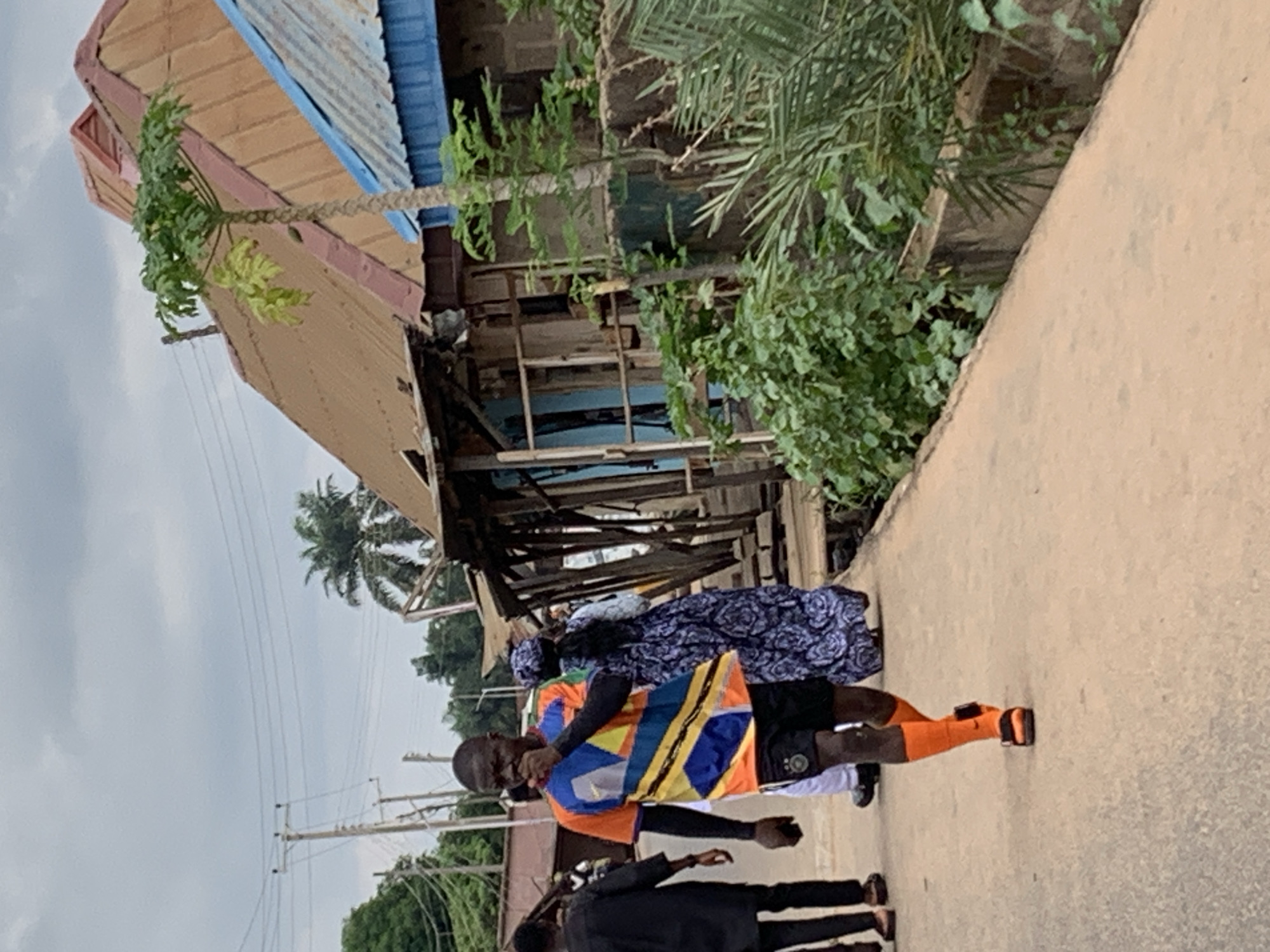 A street scene in Akwanga, Nigeria. A group of people are walking on a concrete path. One person wears an orange, blue, yellow, and black shirt, black shorts, and orange socks with black shoes. Overhead, a dilapidated building has a corrugated metal roof, a partially visible blue portion, and wooden support beams. A palm tree sprouts from the building's structure. A green, overgrown area lines the path. A cloudy sky is above. Electrical lines are visible.