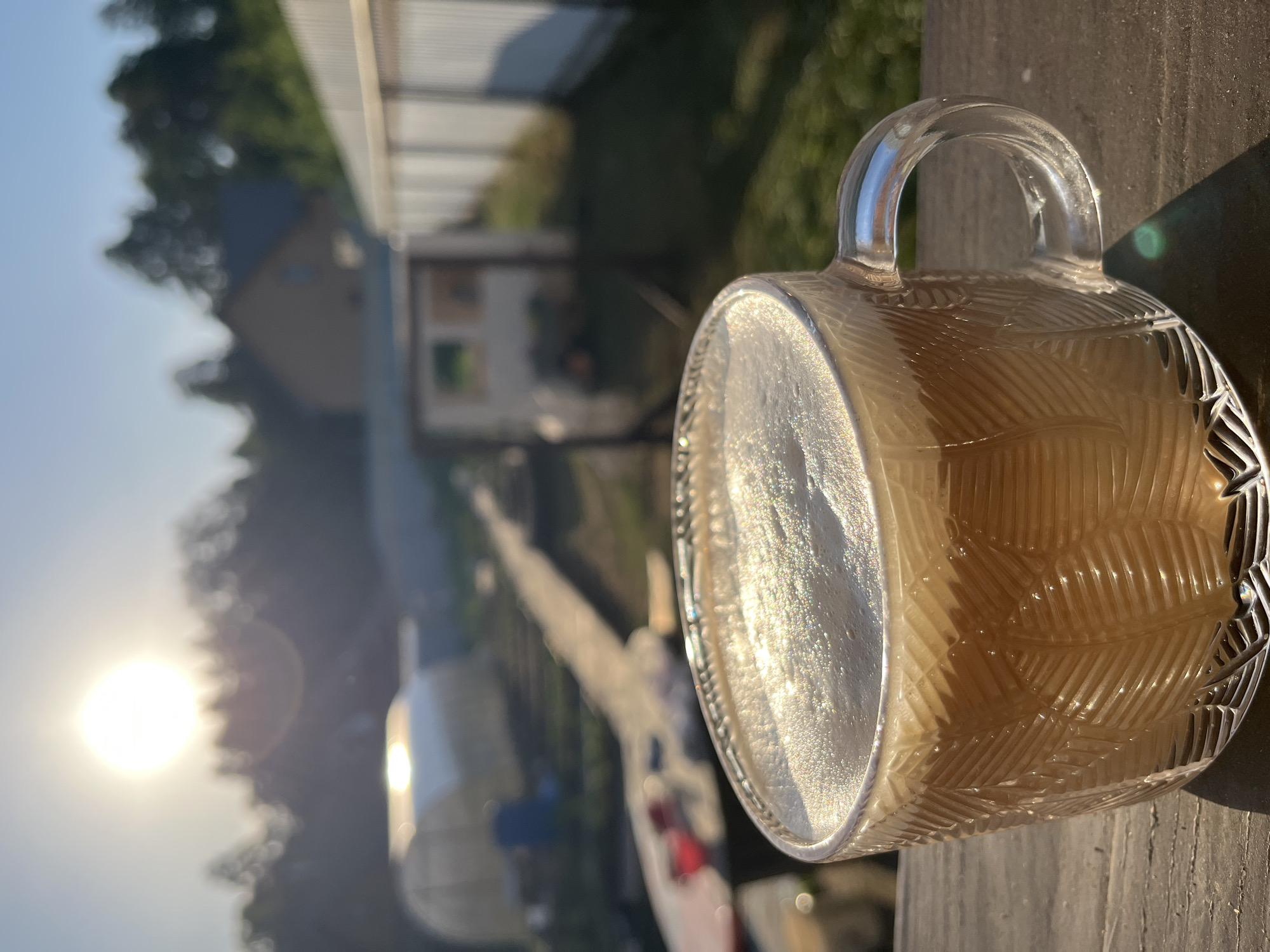 The image shows a clear glass mug with a handle, filled with a light-colored beverage that has a frothy top. The mug sits on a wooden surface, likely a deck or railing. The background is out of focus, but includes elements of a residential area, possibly in the city of Chaikovsky, Russia. There is a fence, some greenery, a building, and a bright sun, suggesting it's daytime. The photograph captures a casual, outdoor moment.