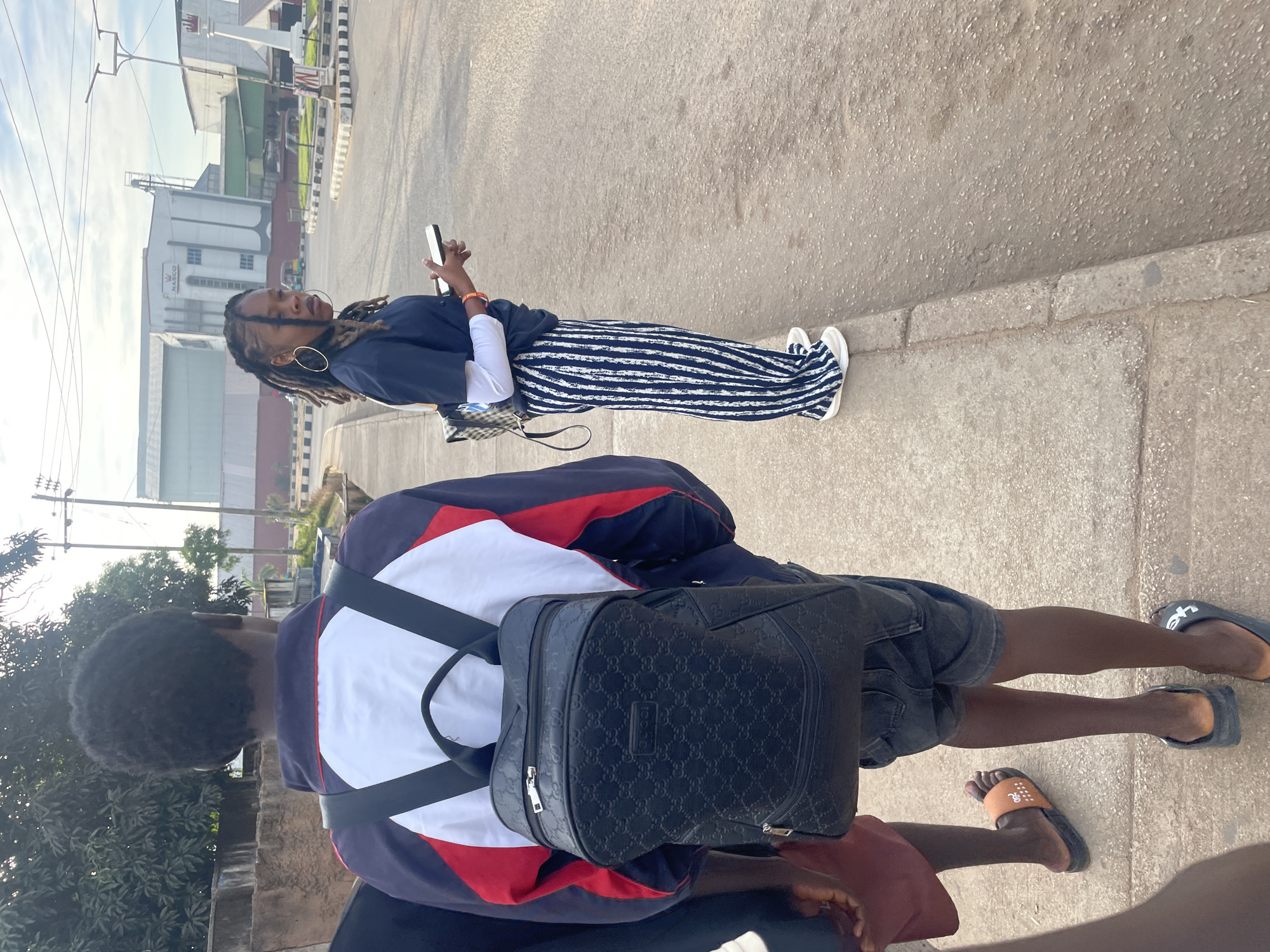 The image shows two young people on a sidewalk next to a road in Jos, Nigeria. The person in the foreground, facing away from the camera, is wearing a jacket with red, white, and blue panels and a black backpack with a repeating pattern. They are also wearing dark shorts and black flip-flops. To the right, a young woman is standing facing the camera and looking to the side. She has dreadlocks and is wearing large hoop earrings. Her outfit consists of a dark blue t-shirt over a white long-sleeved shirt, and wide-legged striped pants. She is holding a smartphone in her right hand. Behind them, there is a road with a roundabout, and in the background, several industrial buildings, including one with a "NASCO" sign. The sky is partly cloudy.