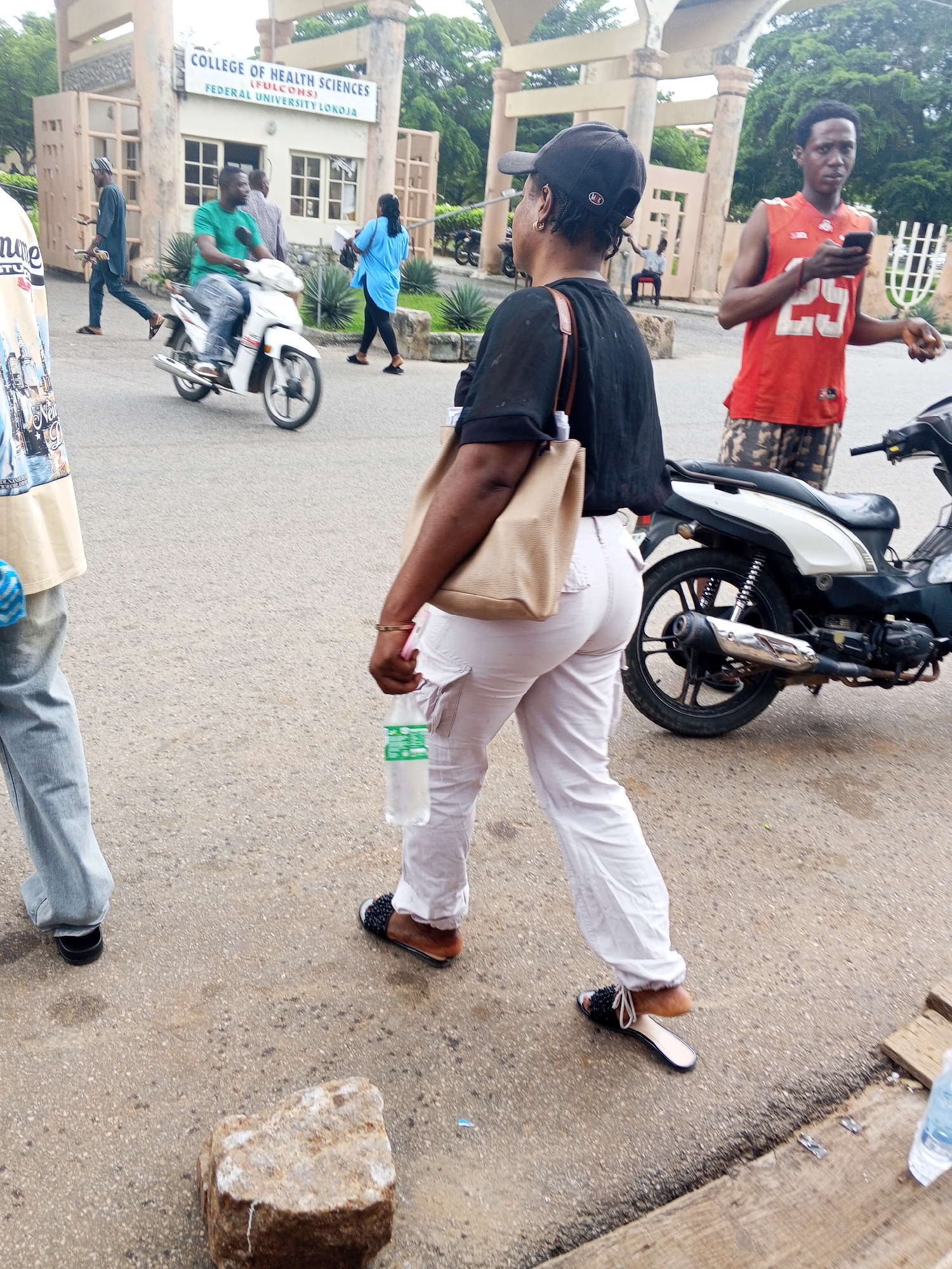 In front of the "COLLEGE OF HEALTH SCIENCES (FULCONS) FEDERAL UNIVERSITY LOKOJA," a woman in a black top and light-colored cargo pants walks away from the camera, carrying a tan purse and a clear plastic bottle of water. She wears dark slides with white straps. To her right, a man in an orange tank top with the number "25" and patterned shorts holds a smartphone. A black and white motorcycle is parked behind him. To the left, a man wearing a green t-shirt rides a white motorcycle. Further left, another person in a blue top walks away. The scene appears to be on a paved road in front of a building with an archway entrance.