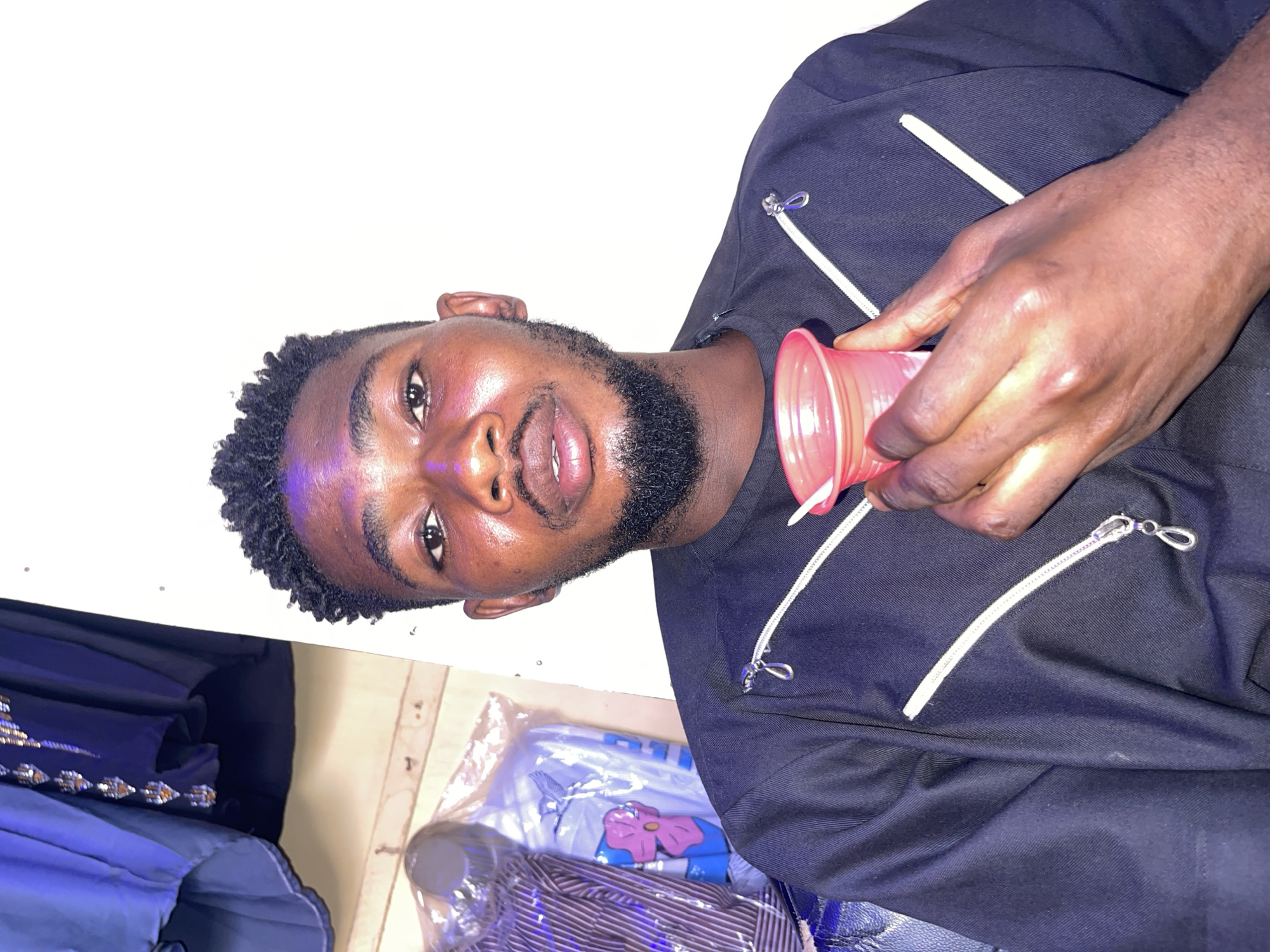 A man with short, dark hair and a beard is holding a pink plastic cup in his right hand. He is wearing a black jacket with white zipper details. The scene includes packaged clothing items, one with a floral design, and a blue garment with silver embellishments. The location is likely an indoor space in Samaru, Nigeria, based on contextual information not directly visible in the image.