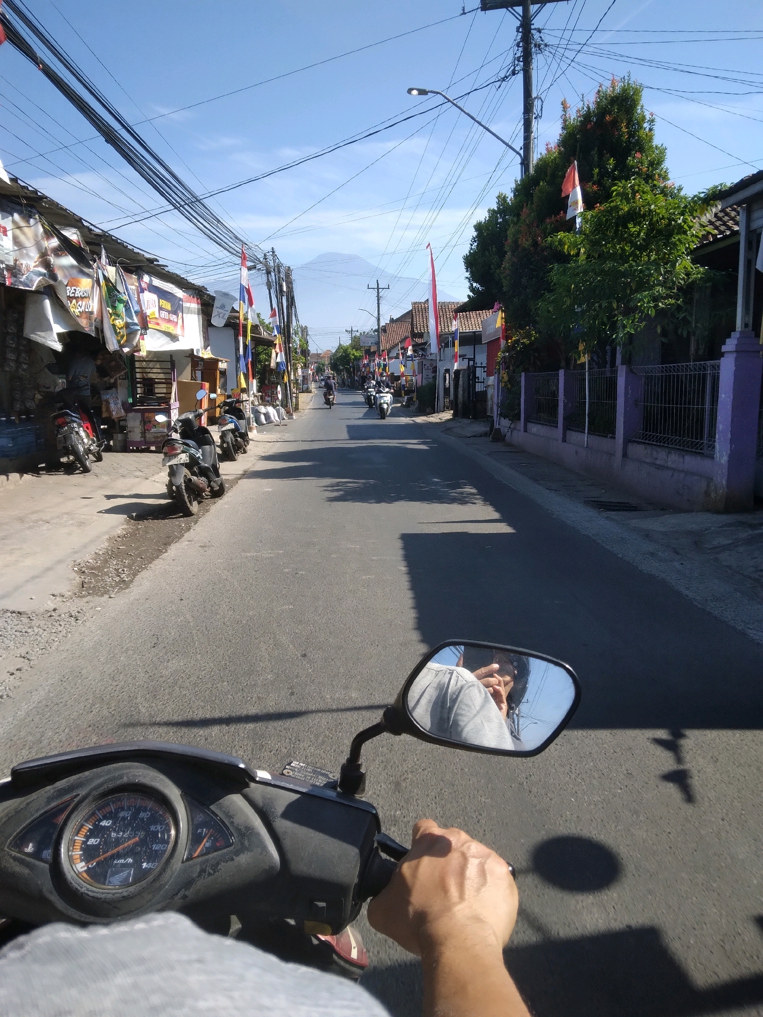 The image depicts a street scene in Purwokerto, Indonesia, viewed from the perspective of someone riding a motorcycle. The road is paved and appears to be a two-lane street. On the left side of the road, there are shops and parked motorcycles. Flags and banners are visible along the street, suggesting a local event or celebration. On the right side of the road, there is a purple fence and lush green trees. The sky is clear and blue.
In the foreground, the dashboard of the motorcycle is visible, including the speedometer. The rider's hand is on the handlebar, and a reflection of the rider in the rearview mirror shows them wearing a gray shirt and a helmet. The rider appears to be taking a photo or video with their phone, as their right hand is raised to their face, holding a phone. Several other motorcycles and riders are visible further down the street, moving away from the viewer. In the distance, a mountain is faintly visible against the horizon, partially obscured by the haze.
