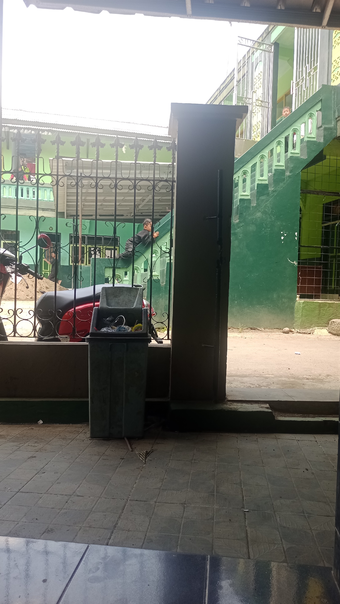 In Karyamandala, Indonesia, an outdoor scene depicts a red scooter parked behind a black metal fence. Next to the scooter is a grey trash can containing various items. A young boy in a black outfit is seen climbing on a green structure, possibly a slide or a staircase. In the background, a green building with white window frames is visible. The ground is paved with grey tiles.