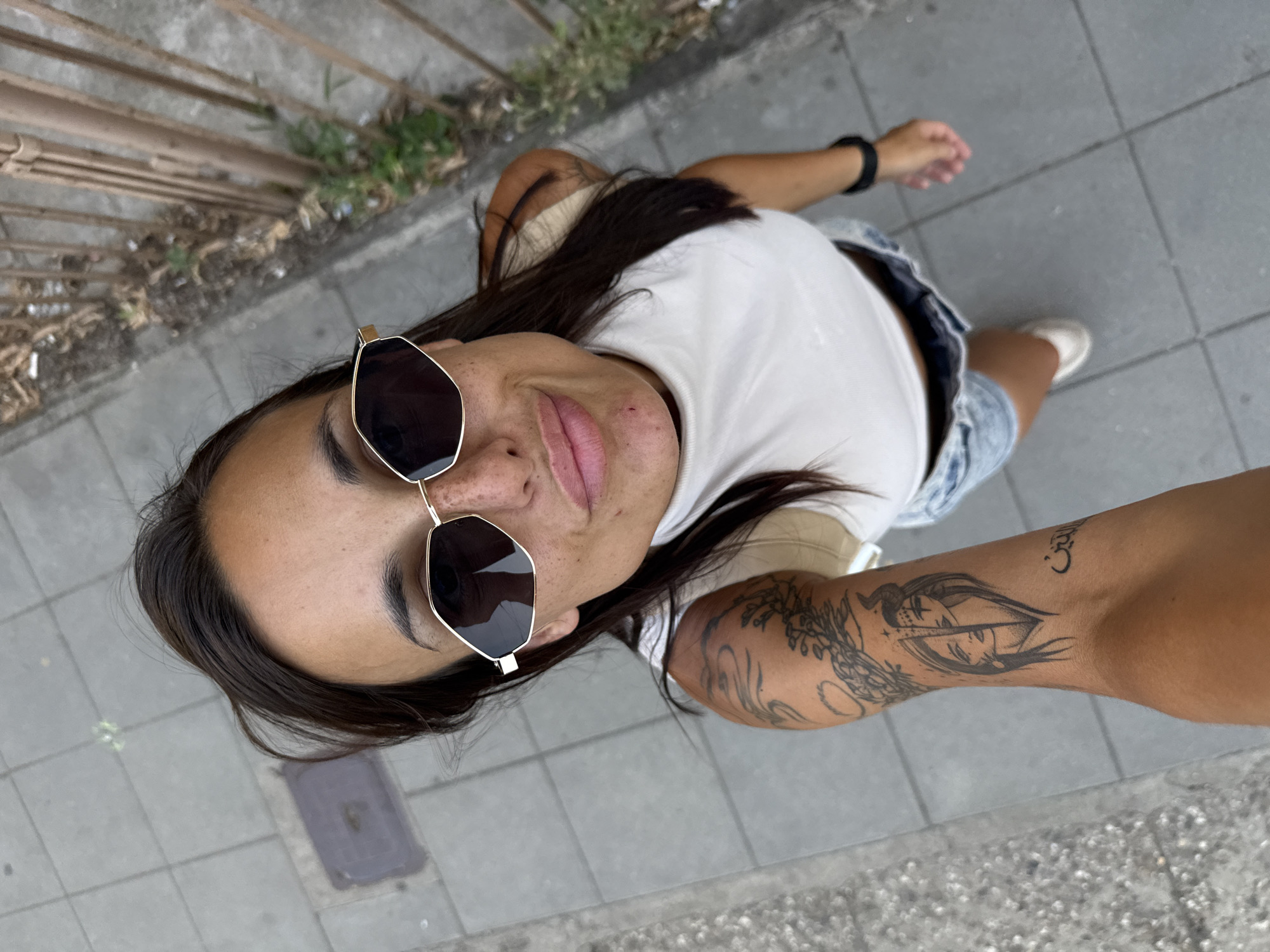 A high-angle medium shot captures a woman with long, dark hair, wearing sunglasses, a white tank top, and denim shorts, posing for a selfie on a sidewalk. The woman has a tattoo on her left arm, and appears to be in motion. The background reveals a concrete sidewalk with a metal fence to the right. The scene is set in the city of Pleven, Bulgaria.
