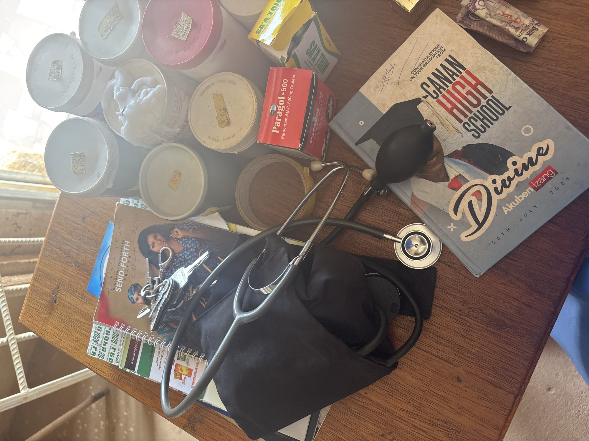 A wooden table in Kuru Karama, Nigeria, displays medical supplies and personal items. There are several white plastic containers, one open revealing cotton inside, a box of "Paragol-500" Paracetamol tablets, and a box of "SAA TRIM." A stethoscope rests atop a black cloth bag. A notebook, keys, and a card titled "SEND-FORTH PRAYER" are also present. Beside the medical items lies a book with "CANAN HIGH SCHOOL" and the name "Divine Akuben Izang, July 26TH, 2025" printed on the cover. Some Nigerian banknotes are nearby. A window and partial view of the exterior is visible.