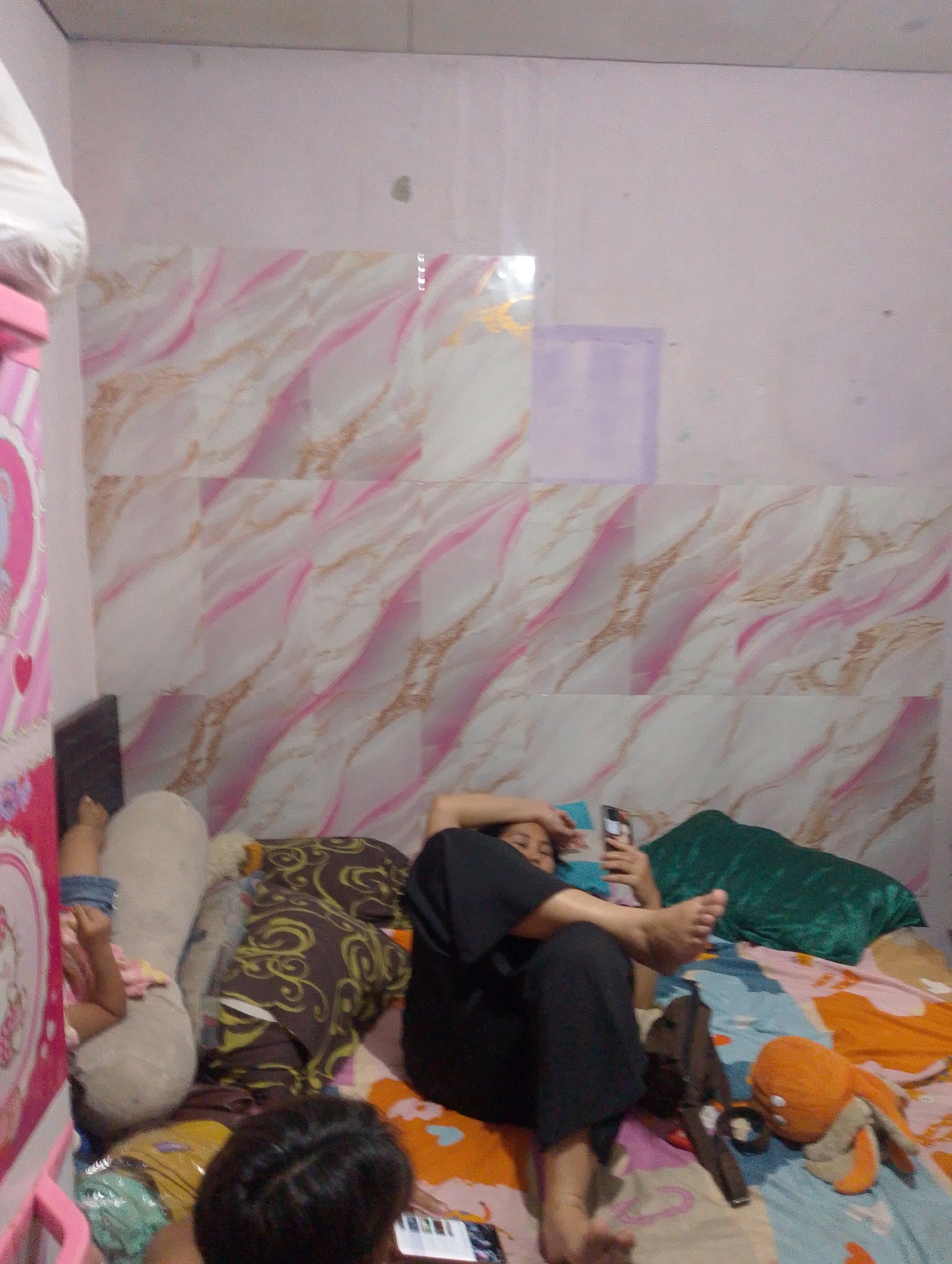 The image depicts an indoor scene, possibly a bedroom, with a person lying on a bed. The bed is covered with a colorful patterned duvet. A person is seen lying on their back, looking at a mobile phone. To the left of the bed, another person's legs are visible, positioned as if they are also lying down. The wall behind the bed has a patterned wallpaper with pink and gold streaks. Various objects like pillows, a stuffed toy, and a bag are scattered on the bed. The room appears to be dimly lit. The context of South Jakarta, Indonesia, suggests a residential setting.