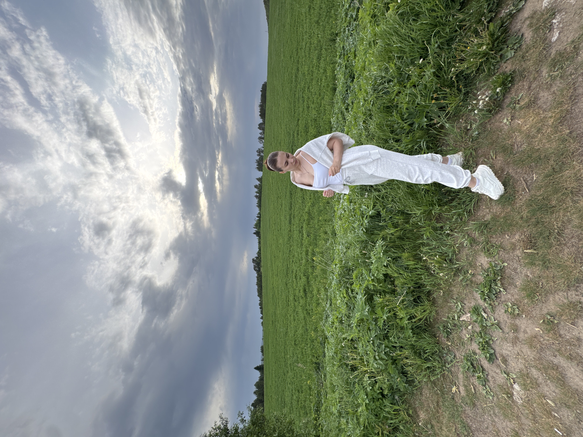 A person wearing a white outfit stands on a path next to a field under a cloudy sky. The person appears to be female and is wearing white sneakers. The field is green and stretches to the horizon. The sky is filled with gray and white clouds. The location is likely rural and possibly near Lukintsy, Russia.