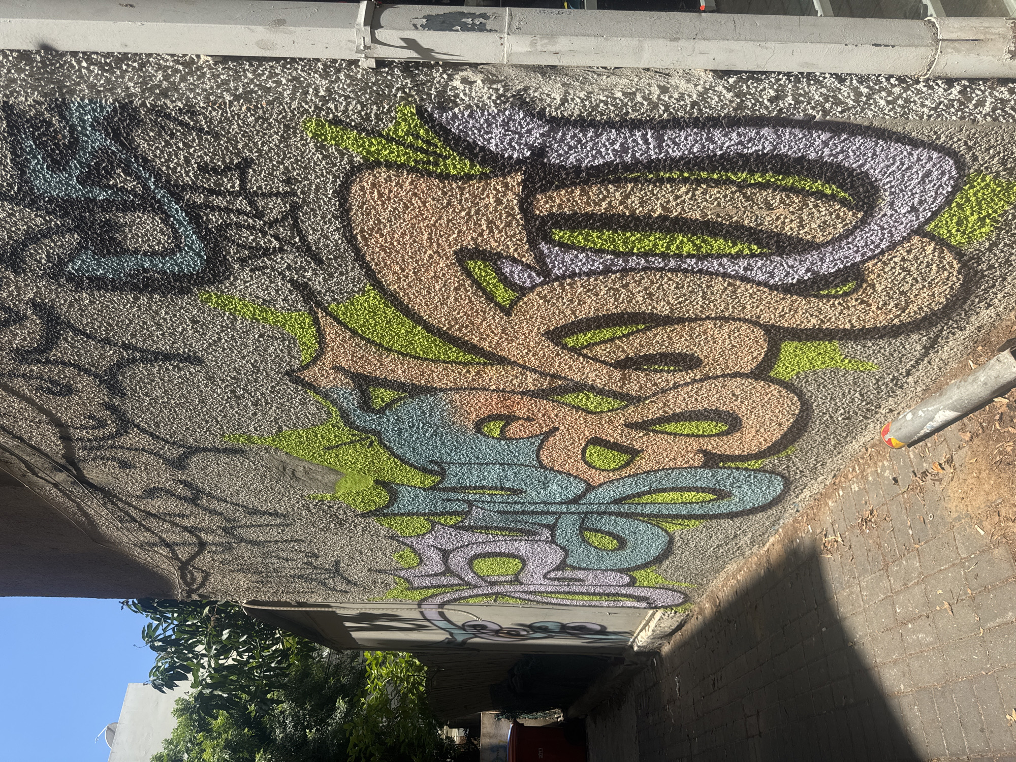 The image shows a concrete wall covered in colorful graffiti, located in Tel-Aviv, Israel. The graffiti features abstract shapes and lines in colors such as beige, purple, blue, and green. A metal pipe runs along the top of the wall, and part of a sidewalk is visible on the right side. Below the wall, there is greenery and a glimpse of a blue sky. The scene is bathed in sunlight.