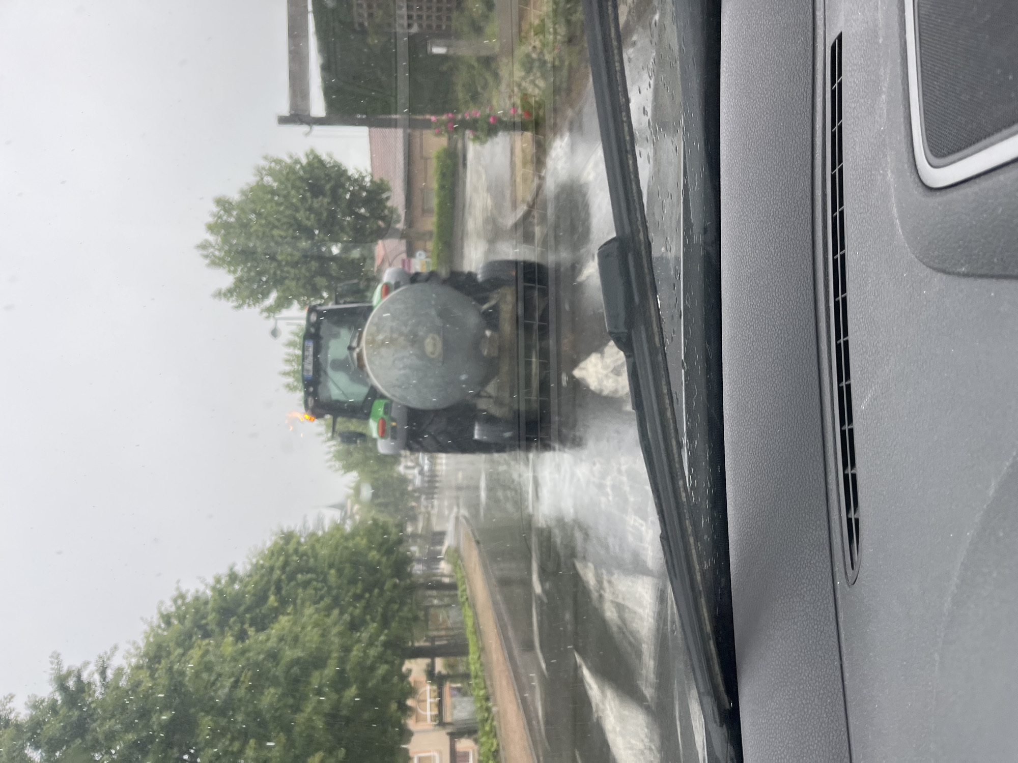 The image, taken from inside a vehicle, shows a tractor with a tank driving on a wet road in Chambœuf, France. Trees line the street, and buildings are visible in the background. The windshield wipers are in motion.