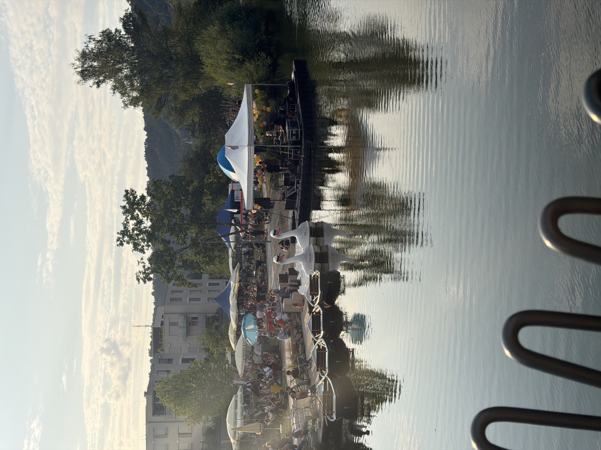 This is a wide-angle, eye-level, outdoor shot taken on a sunny day with some clouds in the sky. The scene depicts a waterfront area with a body of water in the foreground and a busy embankment in the background. Several small boats, some shaped like swans, are docked near the shore.
On the embankment, a gathering of people is visible, seated under various umbrellas and tents. A white tent with a blue dome serves as a stage for a band performing on a floating platform. Colorful balloons are strung around the tent. Behind the embankment, apartment buildings and trees are seen, suggesting an urban setting. The reflection of the sky, trees, and buildings is visible on the rippling water. The image appears to be taken in Eichstätt, Germany, given the context of a waterfront event with a band and recreational boats.
