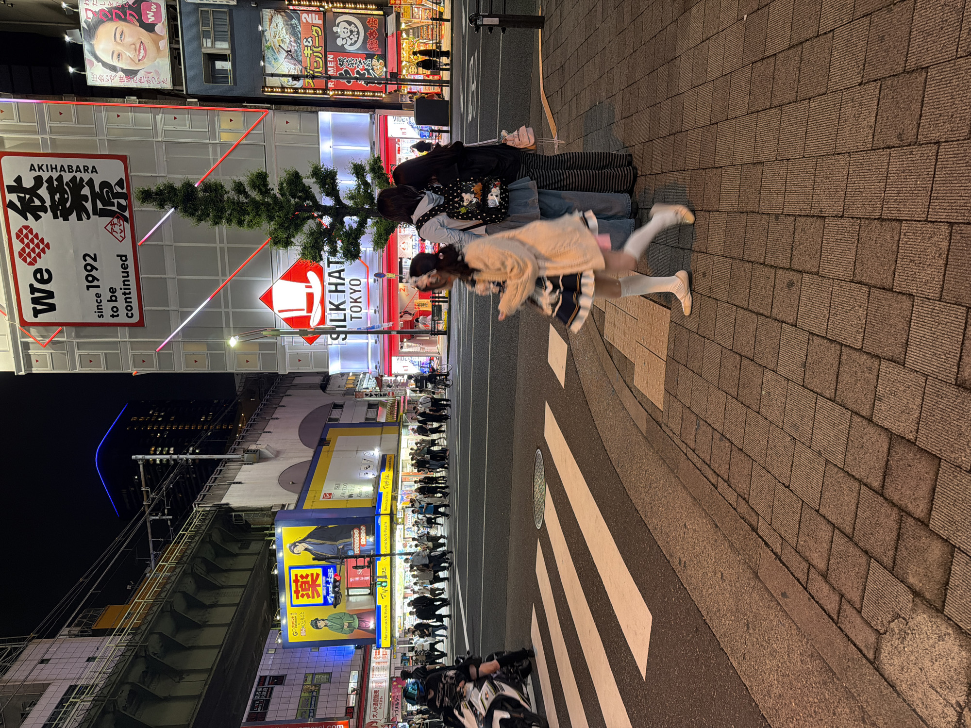 In Chiyoda, Japan, at night, a street scene unfolds with a pedestrian crossing. A person on a white motorcycle is on the far left. The street features a zebra crossing with white stripes. The sidewalk is paved with brown rectangular tiles. Several pedestrians are crossing the street in the background, illuminated by bright neon signs. One prominent sign displays "We❤秋葉原 AKIHABARA since 1992 to be continued". Another sign features a cartoon image of a white hat and the text "SILK HAT TOKYO". A tall evergreen tree stands near the Silk Hat sign. Three people are standing on the sidewalk on the right side of the image, with one person in the foreground walking across the zebra crossing. This person is wearing a light-colored cardigan over a dark dress with white accents, white knee-high socks, and light-colored ballet flats. Buildings with glowing signs and lights line the street. An elevated railway track is visible in the upper left portion of the image.