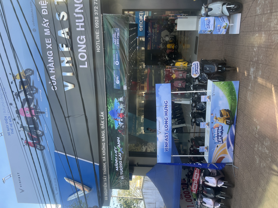 The image displays the exterior of a VinFast electric motorcycle dealership, specifically identified as "VINFAST LONG HƯNG," situated at 69 Nguyen Tat Thanh, Krong Nang, Dak Lak Province, Vietnam. The building features a multi-level facade under clear daylight. The uppermost blue banner advertises "XE MÁY ĐIỆN THẾ HỆ MỚI" (New Generation Electric Motorcycles) and showcases five distinct VinFast electric scooter models: Vento S, Theon S, Feliz S, Klara S, and Evo 200. Below this, a large grey sign prominently displays the VinFast logo and the store name "VINFAST LONG HƯNG," with the full address "69 NGUYEN TAT THANH, XA KRONG NANG, ĐẮK LẮK" and a hotline number "0928 372 777" visible. A green banner beneath advertises promotions, including "MẠNH LIỆT VÌ TƯƠNG LAI XANH" (Strong for a Green Future), a 6% interest rate, 0 down payment, and free battery charging/swapping. Numerous utility lines crisscross the sky above the signage.
On the tiled pavement in the foreground, a promotional setup is active. To the left, a blue pop-up canopy covers several VinFast electric scooters, predominantly in light purple/pink and white, with a price display stating "13.500.000". In the center, a blue and white promotional booth for "VINFAST LONG HƯNG" displays a graphic of a yellow scooter and the slogan "ĐỔI XĂNG LẤY ĐIỆN" (Swap gasoline for electricity), with several brochures placed on its counter. A black VinFast scooter is positioned directly in front of this booth. Additional dark-colored VinFast scooters are parked in rows behind the booth. To the far right, a white VinFast scooter is presented next to a stack of blue promotional boxes.
Through the large glass entrance of the store, numerous VinFast electric scooters of various colors, including yellow, red, black, pink, white, and green, are visibly displayed on a polished interior floor. One male individual is partially visible standing inside the store in the background, near the scooters.