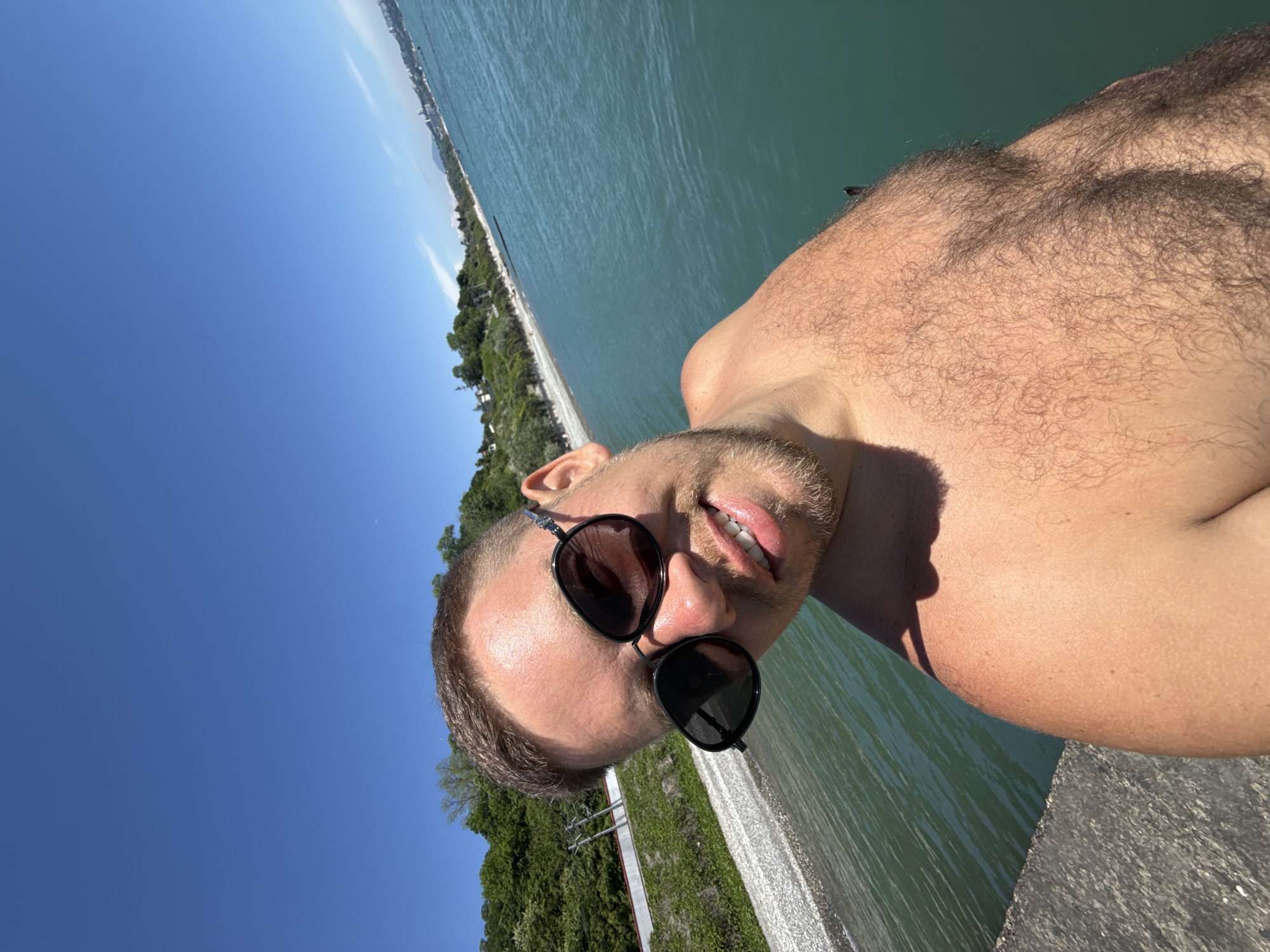 A man takes a selfie outdoors. He wears sunglasses. The man is positioned near a body of water, likely a lake or sea. The background features a shoreline with trees and buildings in the distance. The setting suggests a sunny day, with a clear blue sky above. Given the mention of Sochi, Russia, it is plausible this location is on the Black Sea coast.