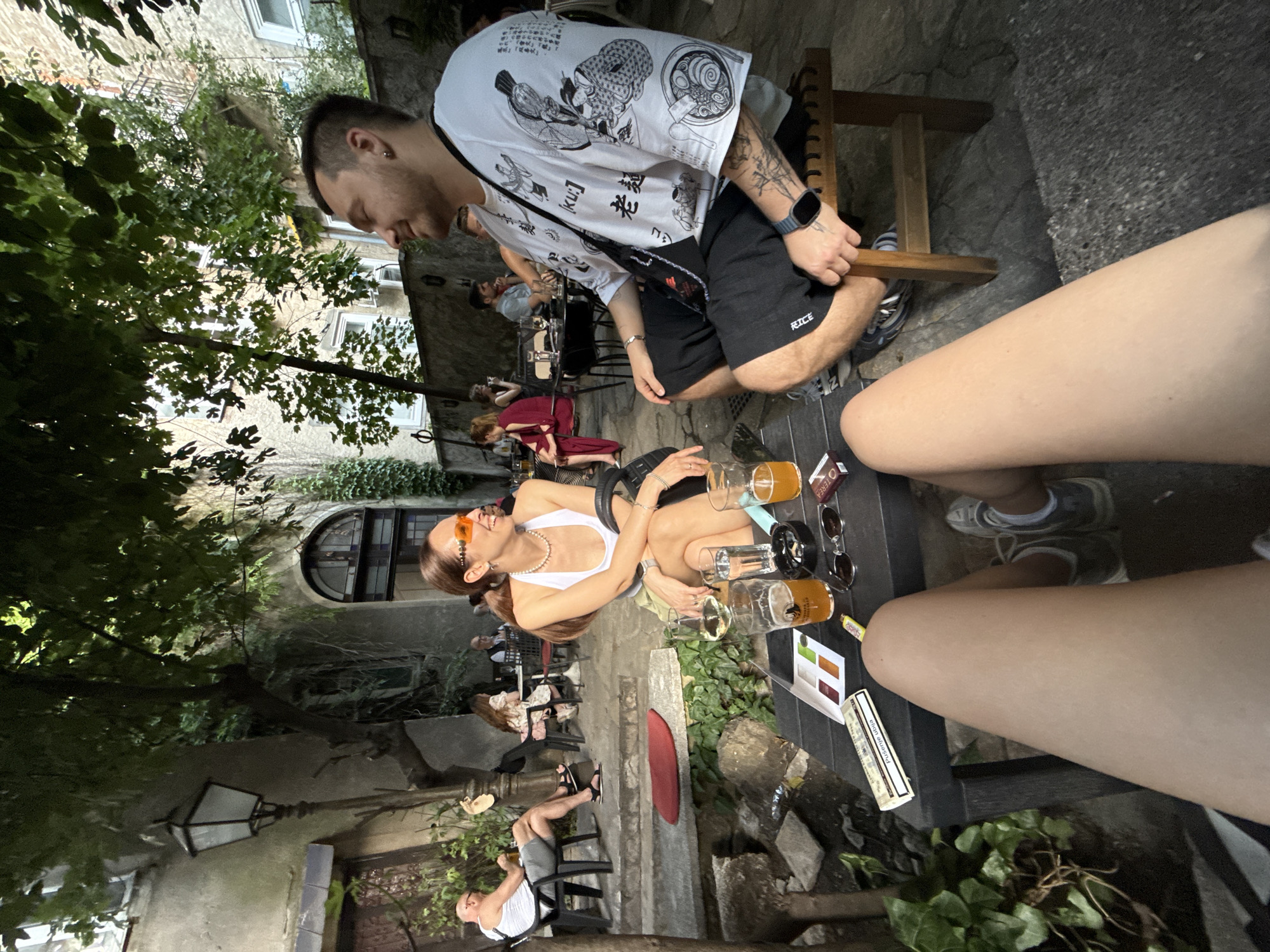 In an outdoor courtyard setting that appears to be located in the City of Zagreb, Croatia, a group of people are seated around tables, seemingly at a cafe or restaurant. In the foreground, a person's legs are visible, and a table in front of them holds several items including glasses of drinks, a pack of cigarettes, and sunglasses. To the right of the table, a man wearing a white t-shirt with Japanese-style graphics and black shorts is seated. He has tattoos on his arms and is looking to his left. Opposite him, a woman with brown hair, wearing a white top and a skirt, and orange-tinted sunglasses, is also seated. She is looking to her left and smiling. In the background, other patrons are visible, some seated at tables, and the courtyard is bordered by buildings with windows and ivy. A lamppost is also present in the scene. The overall atmosphere suggests a casual social gathering.