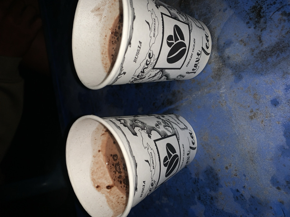 This media file presents a close-up view of two partially consumed disposable paper cups, resting on a textured, dark blue surface, possibly a table or tray. The scene is set indoors, with dim lighting suggesting either evening, night, or an interior with low artificial light, consistent with locations like a cafe or public transport in Metro, Indonesia.
The white paper cups are decorated with intricate black illustrations. These designs include a stylized world map with clearly visible country names such as "FRANCE," "RUSSIA," "MONGOLIA," and "INDIA." The "INDIAN OCEAN" is also distinctly labeled. Each cup features a prominent square logo depicting two stylized coffee beans. Vertically aligned text on the side of the cups plainly reads "Leave Tea."
Inside both cups, there is a brown, frothy residue with settled dark particles, indicating that the beverages, likely hot chocolate or coffee, have been mostly consumed. A dark, blurry shape is visible in the background on the left, possibly a part of a person's limb or another object. The overall image captures a casual moment of refreshment or a break.