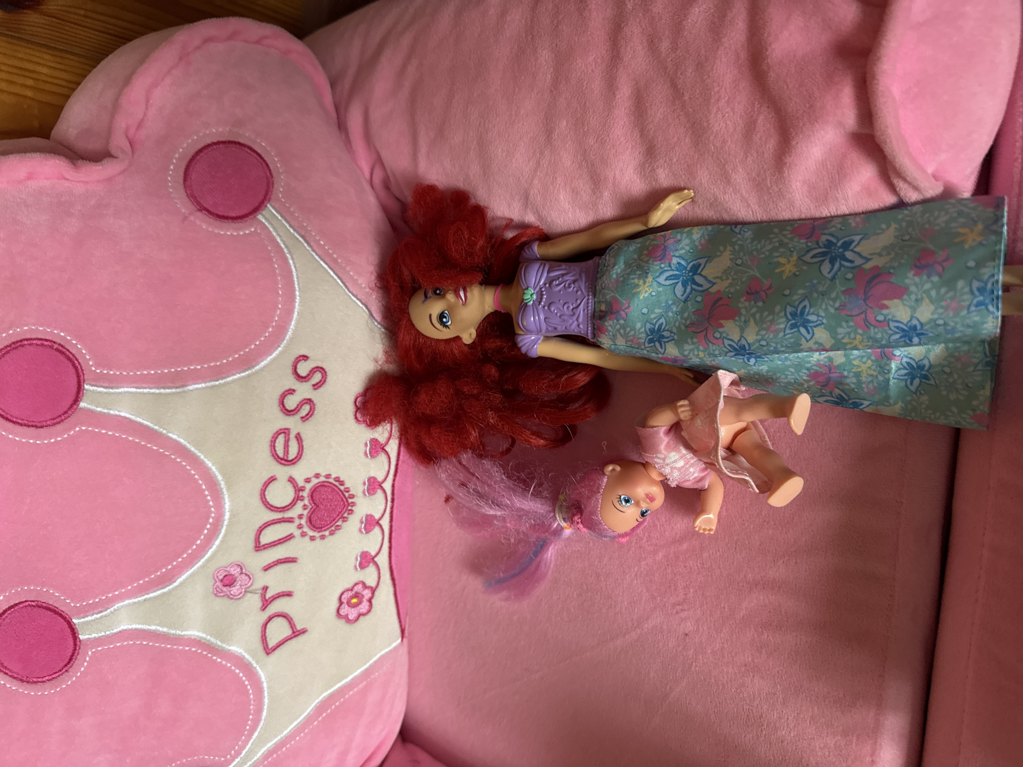 In a close-up, overhead shot, two dolls lie on a plush pink surface, likely a child's bed or play mat. The backdrop is a large, pink, crown-shaped pillow with "Princess" embroidered in pink cursive script. To the left, a smaller doll with bright pink hair, blue eyes, and a pink dress is positioned as if sitting up, with its left arm extended. To the right, a larger doll, resembling Ariel from Disney's "The Little Mermaid," with fiery red hair, a purple top, and a blue and floral print skirt, lies on its back. The lighting is bright, casting a slight shadow to the right of the dolls, indicating the light source is overhead and slightly to the left. The environment is a soft, indoor setting, consistent with a child's bedroom.