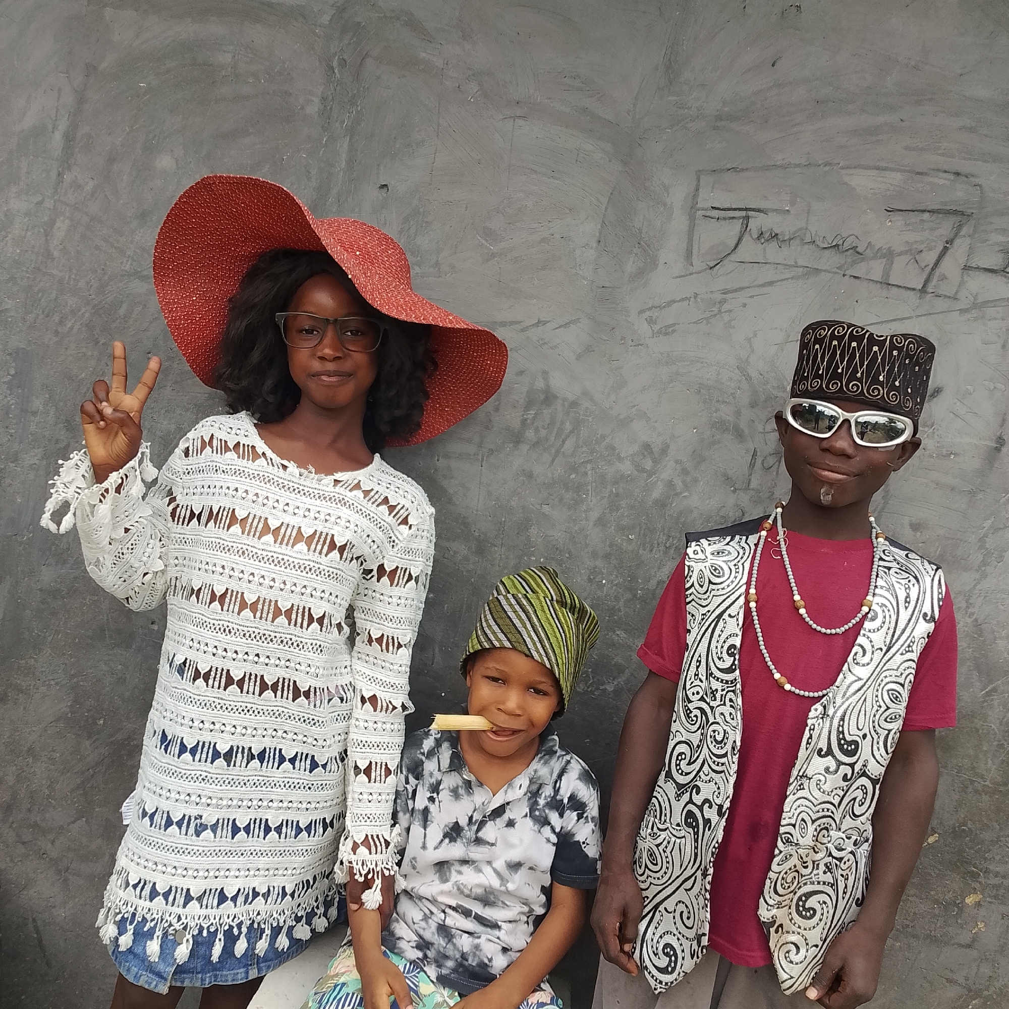 Three young individuals pose in front of a gray, textured wall in Ado, Nigeria. On the left, a girl wears a large, red, wide-brimmed hat, clear-rimmed glasses, and a white, lacy, long-sleeved dress over a denim skirt. She is holding up her left hand, forming a peace sign. In the center, a younger child, seated, wears a striped green and black head wrap and a tie-dye t-shirt. A wooden object is held horizontally in their mouth. On the right, a boy wears a patterned black and gold cap, oversized white sunglasses, a red t-shirt, a black and white patterned vest, and a beaded necklace. The wall behind them has faint, dark scribbles. The lighting is even, suggesting an outdoor setting during daylight.