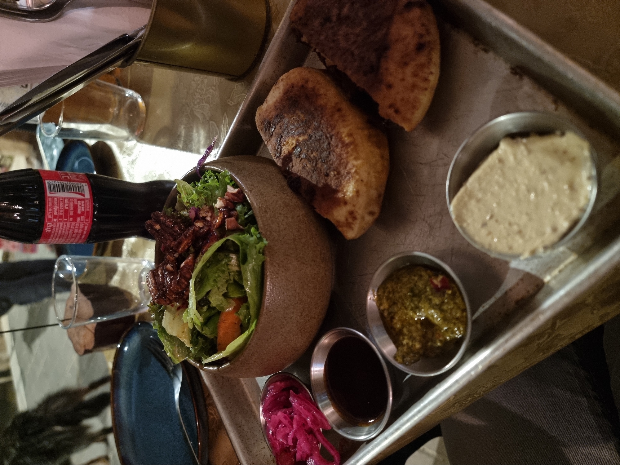 The image shows a food tray containing two pieces of bread, a salad in a brown bowl, and four small metal containers filled with dips. A bottle of Coca-Cola and a drinking glass are also visible. The location is likely in Mitzpe Ramon, Israel, given the visual style and the food served, which is consistent with Middle Eastern cuisine. No people are directly visible, but the presence of the food and drink suggests a dining setting. The environment appears to be indoors, possibly in a restaurant.