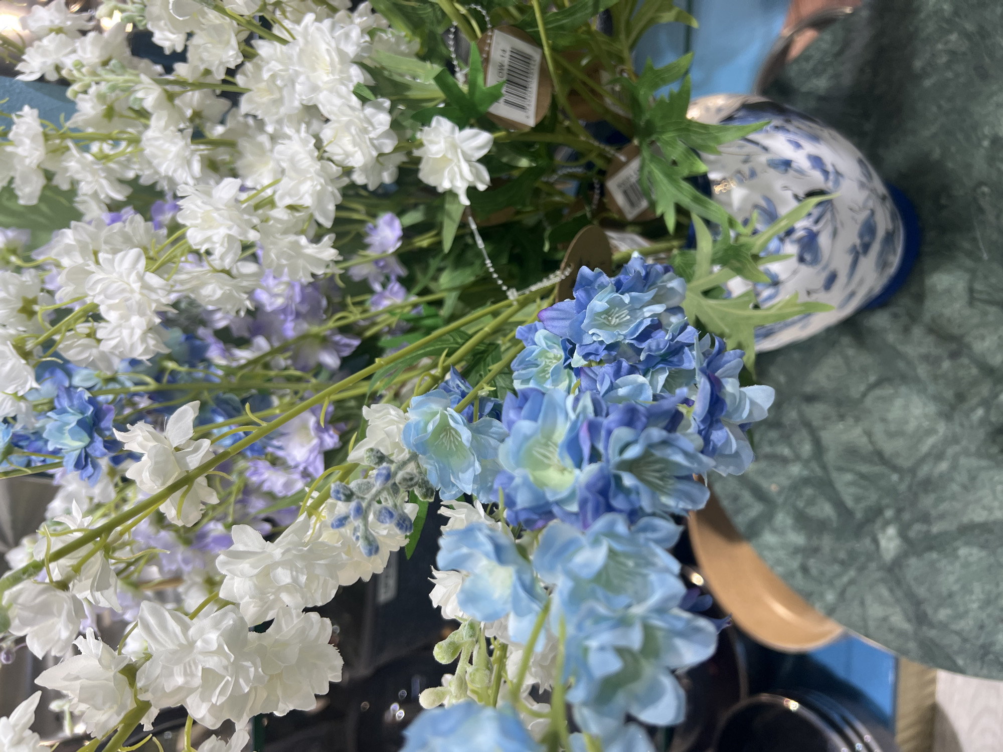 A close-up, eye-level shot captures a bouquet of artificial flowers in a decorative vase. The bouquet features a mix of white, light blue, and lavender flowers, with delicate green leaves interspersed. The flowers are arranged in a tall, slender vase adorned with a blue and white floral pattern. The vase sits on a surface with a dark green, marbled texture. In the background, hints of other objects and surfaces are visible, suggesting an indoor setting. The image is well-lit, with the focus primarily on the flowers and vase. The location in Eshanguzar, Uzbekistan is not discernible from the visual information provided in the image.