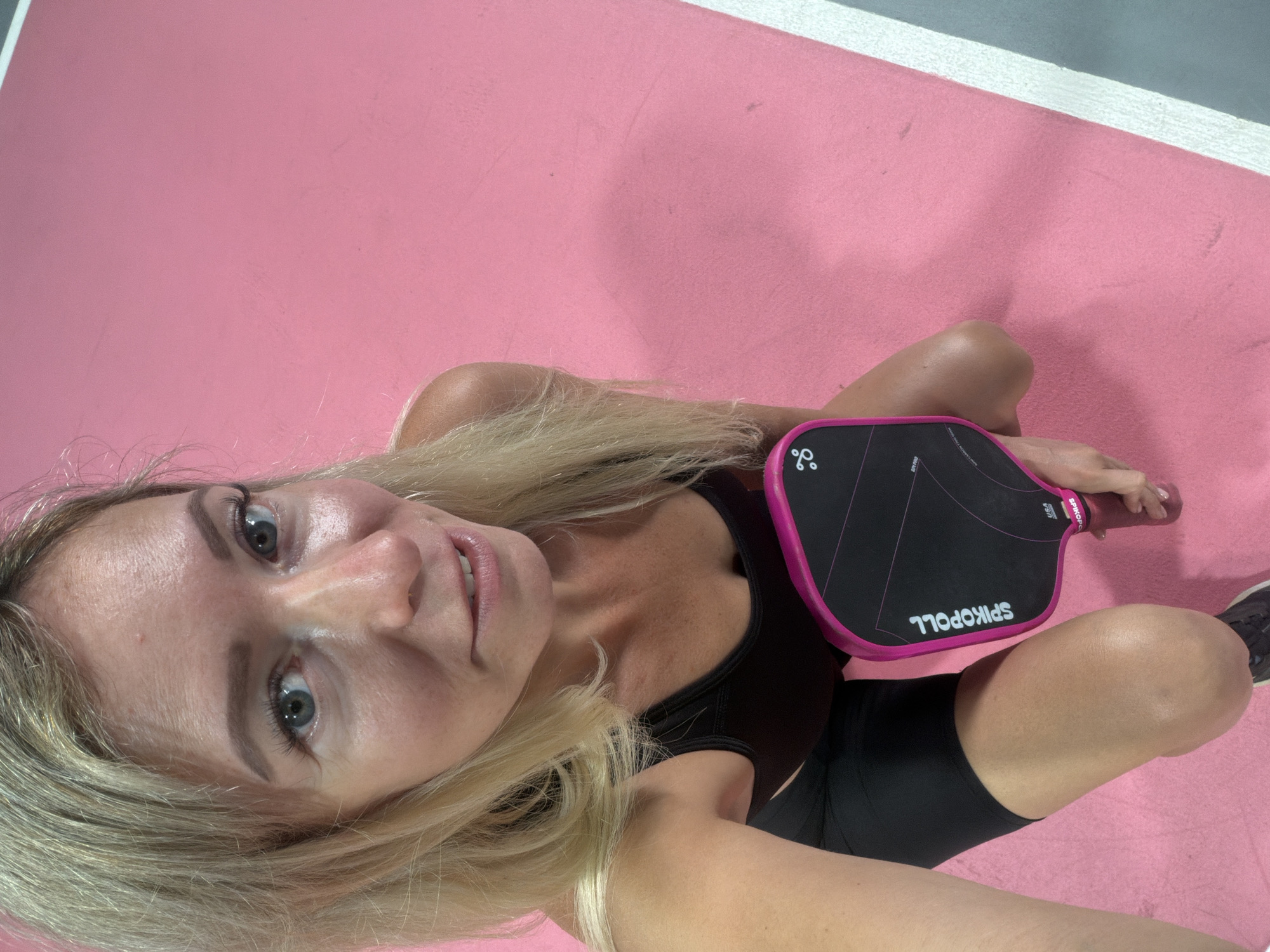 The media file is a vertical, eye-level selfie taken by a blonde woman with blue eyes. She is wearing a black sports bra and black athletic shorts. She is holding a black and pink pickleball paddle in her right hand, which has the brand name "SPIKOPLL" printed on it in white. The woman is positioned on a pink surface, which appears to be a mat or a court. In the background, there is a grey surface with a white line separating it from the pink surface.
The woman is looking directly at the camera with a slight smile. Her blonde hair is long and appears to be slightly windblown. The lighting suggests it is daytime. The overall impression is that of an athletic individual preparing for or taking a break from a game of pickleball. The setting is consistent with an indoor or outdoor sports facility. There is no discernible weather information or time of day cues beyond it being daytime. The emotion conveyed is one of casual enjoyment and readiness for activity.