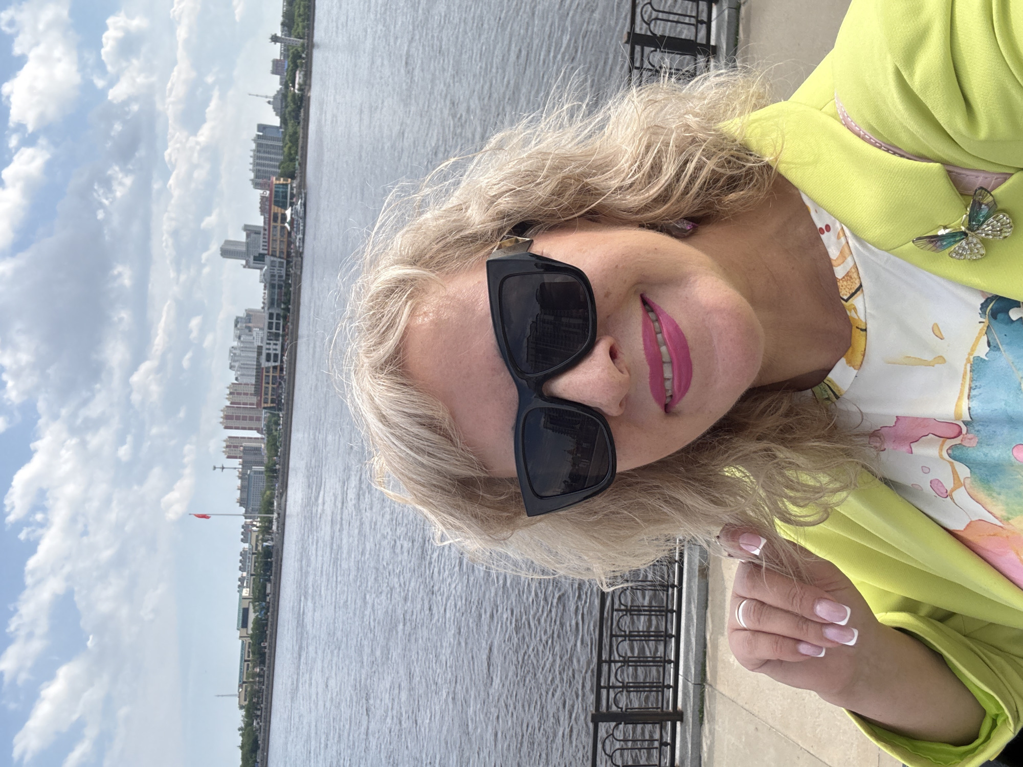 A medium shot photograph captures a woman with blonde, curly hair, wearing large black sunglasses, a bright lime green blazer over a colorful, abstract-patterned top, and a jeweled butterfly brooch. Her lips are painted a vibrant pink, and she is smiling. She is wearing a ring on her left hand, which is resting near her chin. Her fingernails have a French manicure. She is standing on a paved walkway with a metal railing visible behind her. In the background, across a wide body of water, is a city skyline with numerous buildings and structures, including what appears to be a red flag on a pole. The sky is blue with fluffy white clouds. The scene suggests an outdoor setting in the city of Blagoveshchensk, Russia, likely along the Amur River.