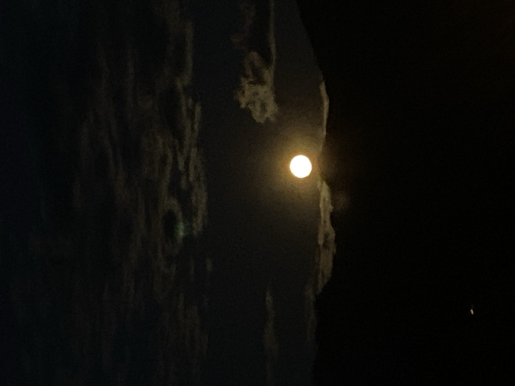 This media file presents a dark, cloudy night sky, likely captured in portrait orientation and displayed horizontally. The central subject is a bright, luminous full moon, positioned towards the right side of the frame. Its intense glow subtly illuminates the edges of the dark, dense clouds that largely obscure it and dominate the left and central portions of the image. These clouds appear as vertical, rugged formations, giving the impression of a darkened, textured landscape or profile against the deep black sky. A faint, tiny point of light is discernible in the bottom right corner of the frame. The scene conveys a sense of serene nighttime natural beauty and mystery. The time of day is night, and the weather is cloudy. No human subjects, specific objects beyond the celestial bodies and clouds, or discernible text are present. The location context provided is Sotonići, Montenegro, though no specific geographical landmarks are visible within the image itself.