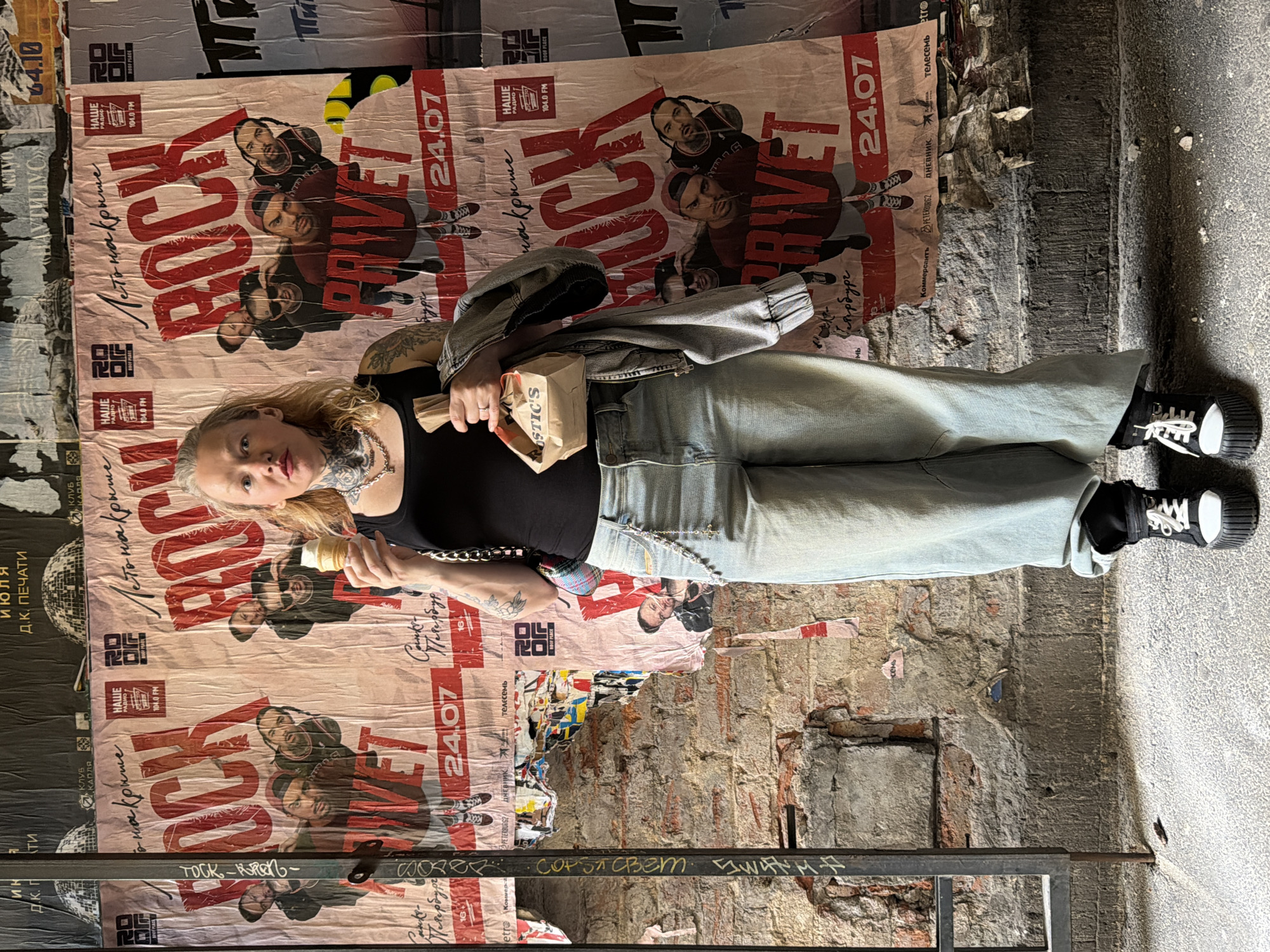 A blonde woman with multiple neck tattoos stands in front of posters in Saint Petersburg, Russia. She is wearing a black tank top, light blue wide-leg jeans, and platform sneakers. She holds a brown paper bag in her right hand and an ice cream cone in her left. The posters advertise a "Rock Privet" event on July 24th, featuring bands and the radio station "Nashi Radio." The background is a brick wall covered in various posters and graffiti.