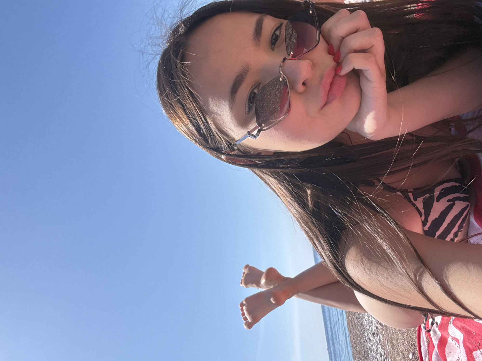 A young woman with long brown hair and sunglasses is posing for a selfie. She is wearing a zebra-striped bikini top and red nail polish. The background is a clear blue sky, a beach with small rocks, and a small portion of the ocean. Her feet are also visible, crossed, indicating she is lying down. The location appears to be a beach. The city and specific location are unknown.