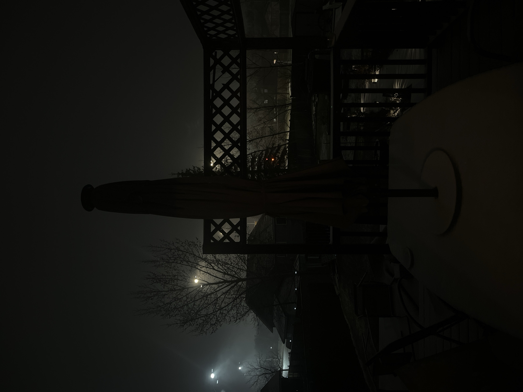 The image shows an outdoor patio scene at night, likely in Calgary, Canada due to the presence of outdoor structures such as a covered gazebo with lattice patterns and outdoor furniture. The environment suggests a suburban or residential area given the street lights and obscured view of buildings in the background. In the foreground, there is a patio table with a closed umbrella. The overall visibility is poor due to the low light and possibly fog or haze in the air. A tree with bare branches is visible in the middle ground, with a street light shining through it. The scene appears still and devoid of people.