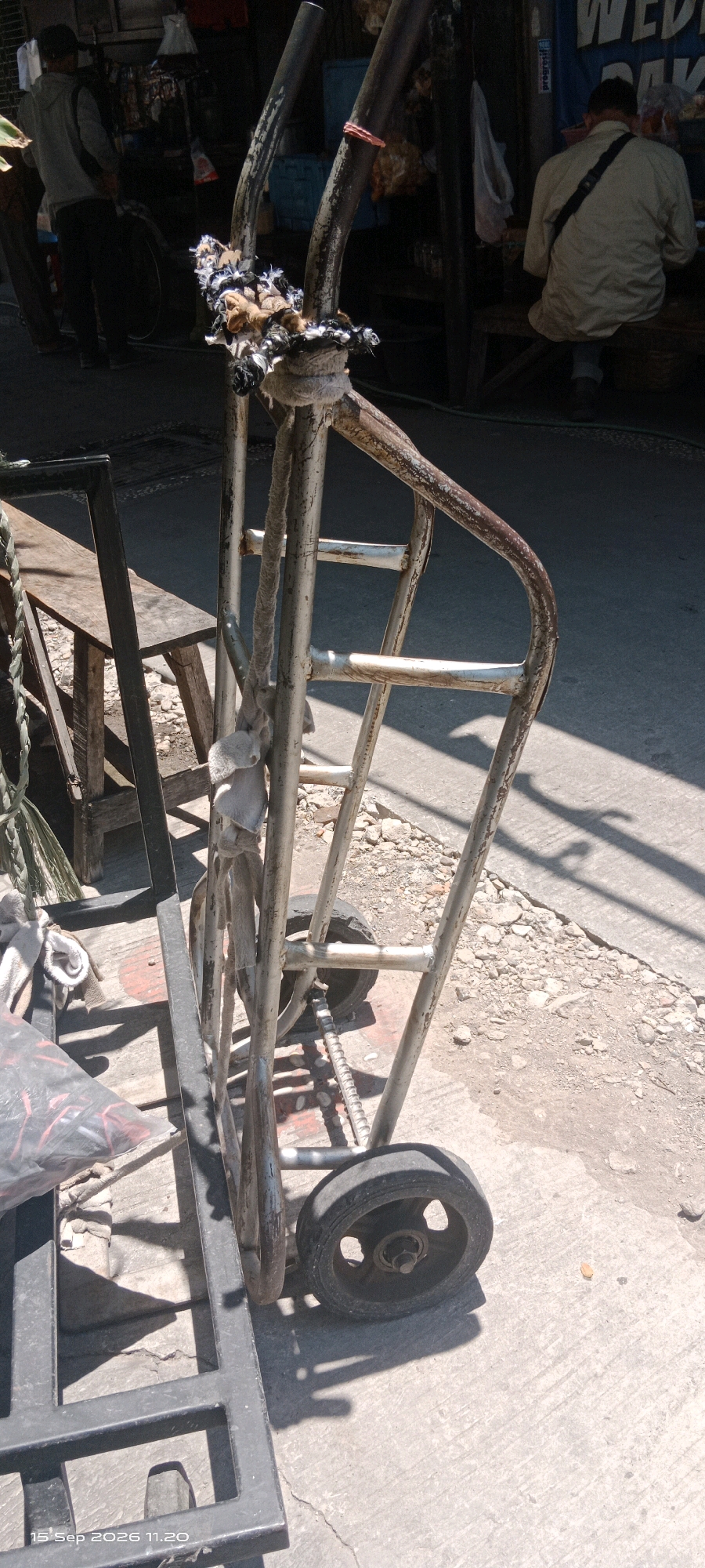 In Surakarta, Indonesia, a close-up shot focuses on the worn metal frame of a trolley. The trolley, which appears to be made of salvaged materials, exhibits signs of rust and wear, with a red elastic band wrapped around one of its uprights. A bundle of frayed rope and fabric is tightly bound to the top of the trolley's frame. A single, worn wheel is visible towards the bottom right of the frame, casting a distinct shadow on the concrete ground. To the left, a portion of a black metal structure, possibly part of another trolley or cart, is visible. In the background, out of focus, two individuals are seen. One person stands with their back to the camera, wearing a gray hoodie and dark pants, while another person is seated. The environment suggests a street or marketplace setting, with shadows indicating bright sunlight.