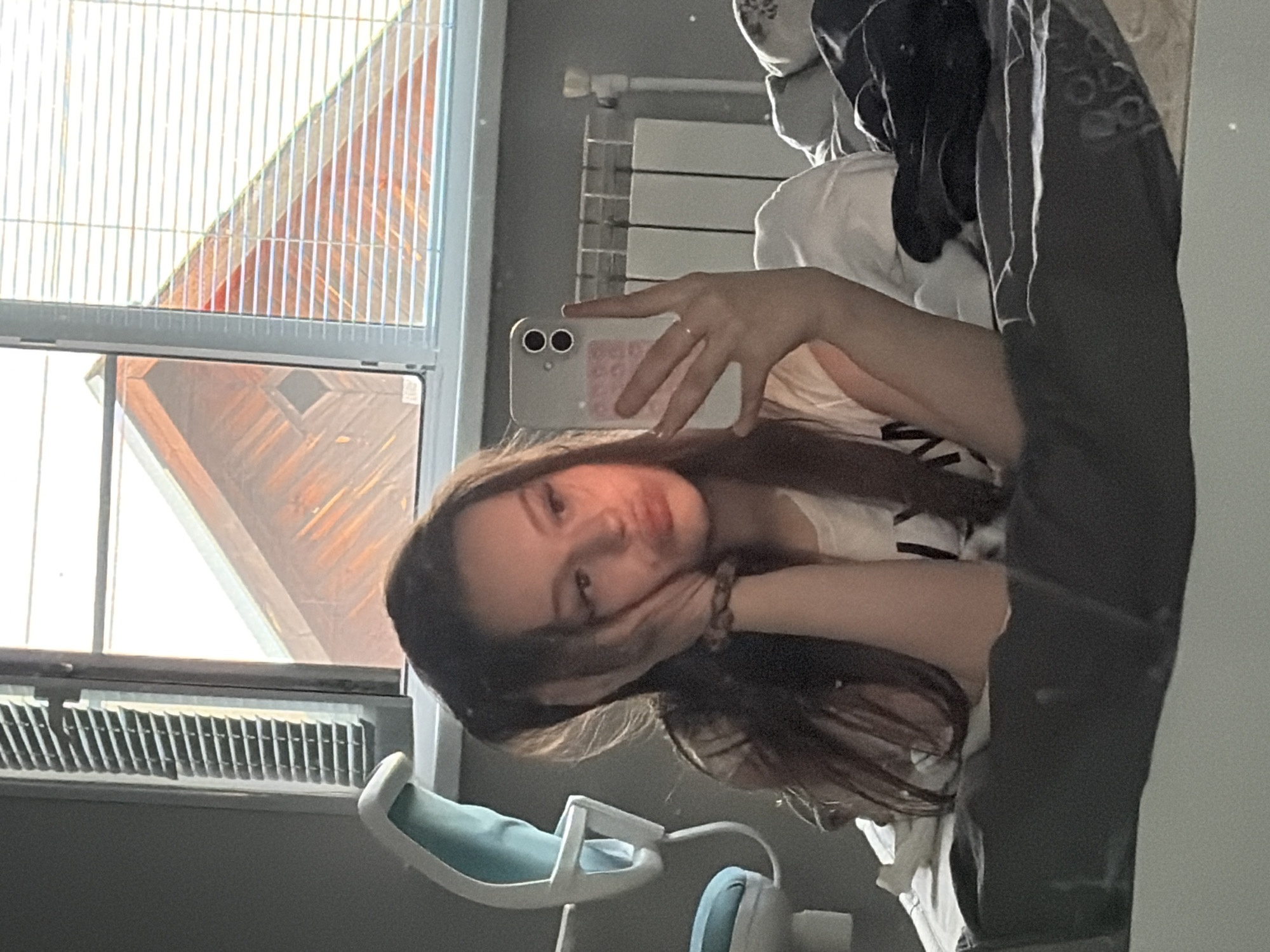 A young female with long, dark hair is positioned on a bed in an indoor setting, identified as Semyonov, Russia. She is taking a mirror selfie, holding a light-colored smartphone with a dual-lens camera to her right. Her left hand rests against her cheek, and her lips are pursed in a pout. She wears a light-colored t-shirt with a visible dark letter and a braided bracelet on her left wrist, with a ring on her right ring finger. The bed is covered with grey bedding. To the immediate left, a light blue and white ergonomic office chair is visible. Behind the subject, a window equipped with venetian blinds reveals an exterior view of a reddish-brown wooden roof featuring a diamond-shaped architectural detail. A white radiator is installed beneath the window on the right side of the frame. The room walls are grey.