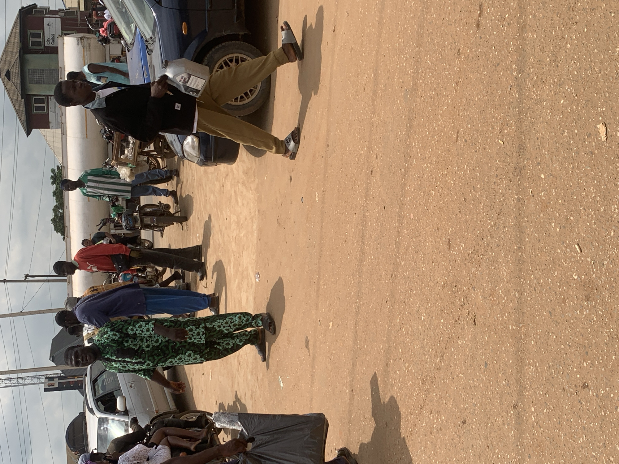 In Shagamu, Nigeria, a scene unfolds on a dusty, unpaved street. A man in a green patterned outfit stands with his hands out, looking directly at the camera, while another man in a black jacket and tan pants walks by carrying a white plastic container. Several motorcycles are parked on the side of the road. A white tank truck with "City Guilds" written on it is visible in the background, along with a red building. The ground is a mix of light brown soil and small pebbles. The sky is overcast, suggesting it might be a cloudy day.