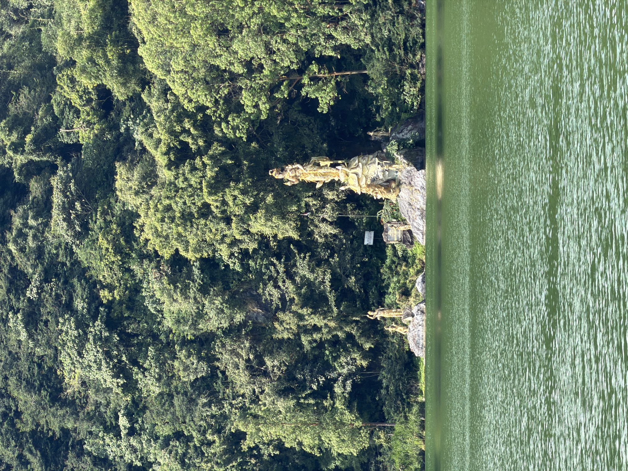 The image shows a body of green water with a textured surface, likely a lake. In the background, a dense forest covers a hillside. On a rocky outcrop near the shore, there are several golden statues. The largest statue is of a multi-armed deity standing on a pedestal decorated with smaller figures. To the left of this central statue, there are two smaller statues of figures in kneeling or seated positions. Further to the right, on a separate rocky area, another smaller golden statue is visible. A white sign is also present on the rocky outcrop. The overall scene depicts a serene natural landscape with religious or artistic sculptures. This location is identified as being in Kintamani, Indonesia.