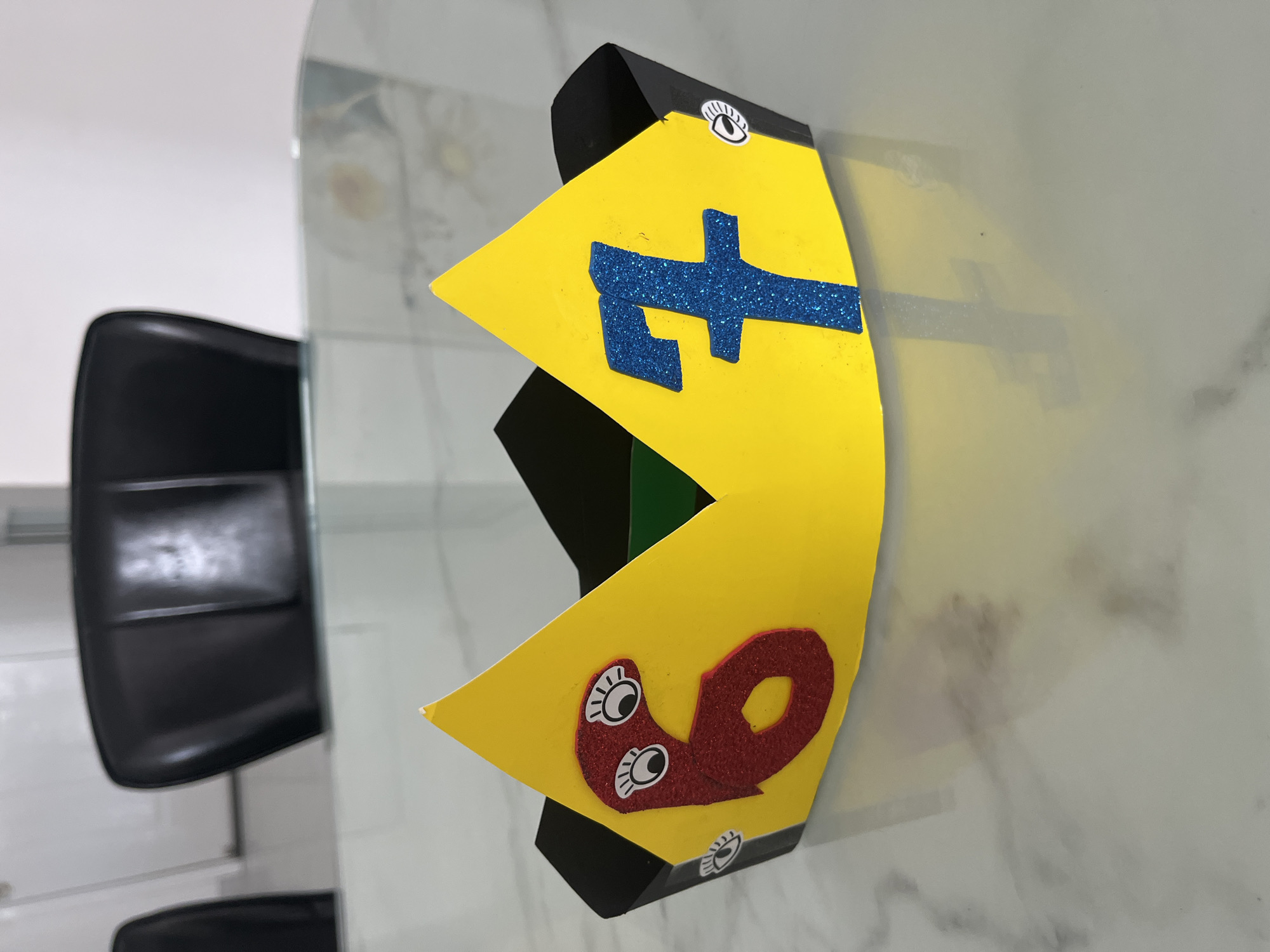 A colorful party crown is lying on a white surface. The crown is made of yellow material and is decorated with a glittery blue "E" symbol and a fuzzy red "9" with cartoon eyes. The crown has black trim on the edges, also decorated with googly eyes. A black chair is visible on the left. The surface appears to be glossy and reflective. Location details are outside the scope of the given image.