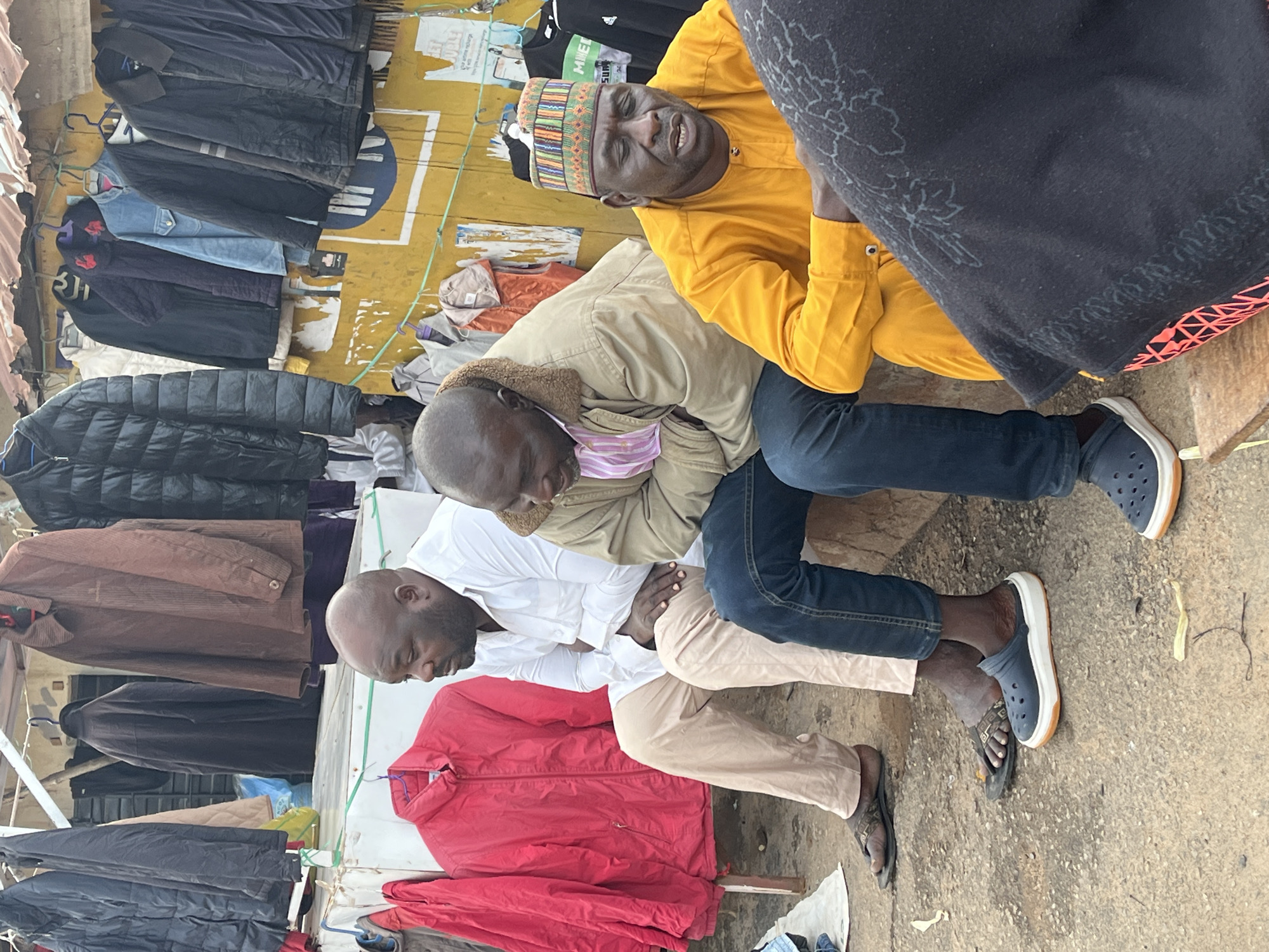 This image captures a street scene in Jos, Nigeria, likely during daytime given the visible lighting. The primary subjects are three men, appearing to be of African descent, engaged in what seems to be a casual interaction or perhaps a business transaction.
The men are positioned in the foreground and mid-ground. The man on the right is wearing a bright yellow garment and a colorful cap, with an expression that suggests he is speaking or laughing. He is seated next to another man wearing a tan jacket over a pink and white striped shirt, who is leaning forward, possibly listening. The third man, in the lower part of the frame, is wearing a white shirt and beige trousers, and is looking down.
The setting appears to be an outdoor market or a street stall. In the background and to the sides, there are numerous garments, mostly jackets and coats, hanging on display. These items are varied in color and style, suggesting a clothing vendor. Some of the garments are dark, such as black jackets, while others are brown and red. The backdrop is a yellow wall with some signage, though the text is mostly illegible or out of focus. One visible sign with white lettering on black is partially obscured but appears to be advertising something.
The ground is paved with concrete and dirt. The overall atmosphere seems relaxed, with the men appearing comfortable in their surroundings. The weather is not explicitly indicated, but the clothing suggests it might not be extremely hot. The presence of hanging clothes and the informal arrangement of people and goods point towards a bustling, everyday scene in a Nigerian urban environment.