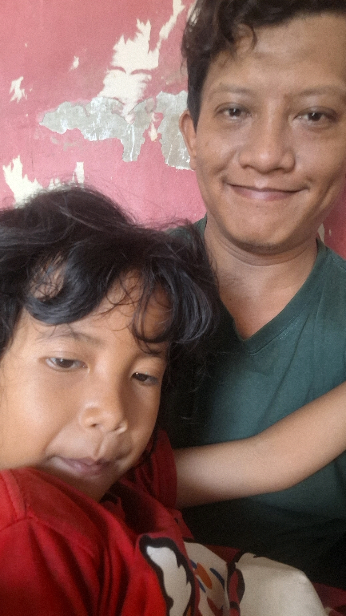 Two individuals, an adult male and a child, are posing for a selfie against a textured pink wall. The adult male, positioned behind and slightly to the right of the child, is smiling and looking towards the camera. He is wearing a dark green v-neck t-shirt. The child, who is in the foreground and positioned to the left, is also looking towards the camera with a slight smile. The child has dark, curly hair and is wearing a red shirt with a graphic design. The background wall shows signs of wear, with peeling paint. The image appears to have been taken indoors or in a sheltered outdoor area.
