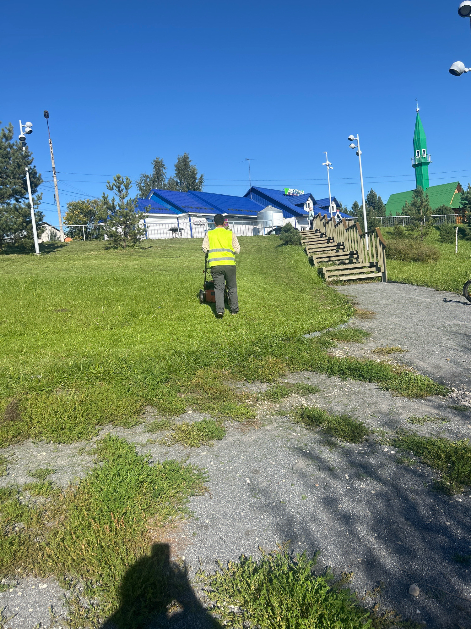 A person wearing a bright yellow safety vest walks away from the camera, pushing a lawnmower on a grassy slope under a clear blue sky. In the background, several buildings with blue roofs are visible, along with a tall green structure resembling a minaret. A wooden staircase ascends the slope to the right of the person. The ground in the foreground and to the right is a gravel path with some green vegetation growing through it. The scene appears to be outdoors, possibly in a park or a public space.