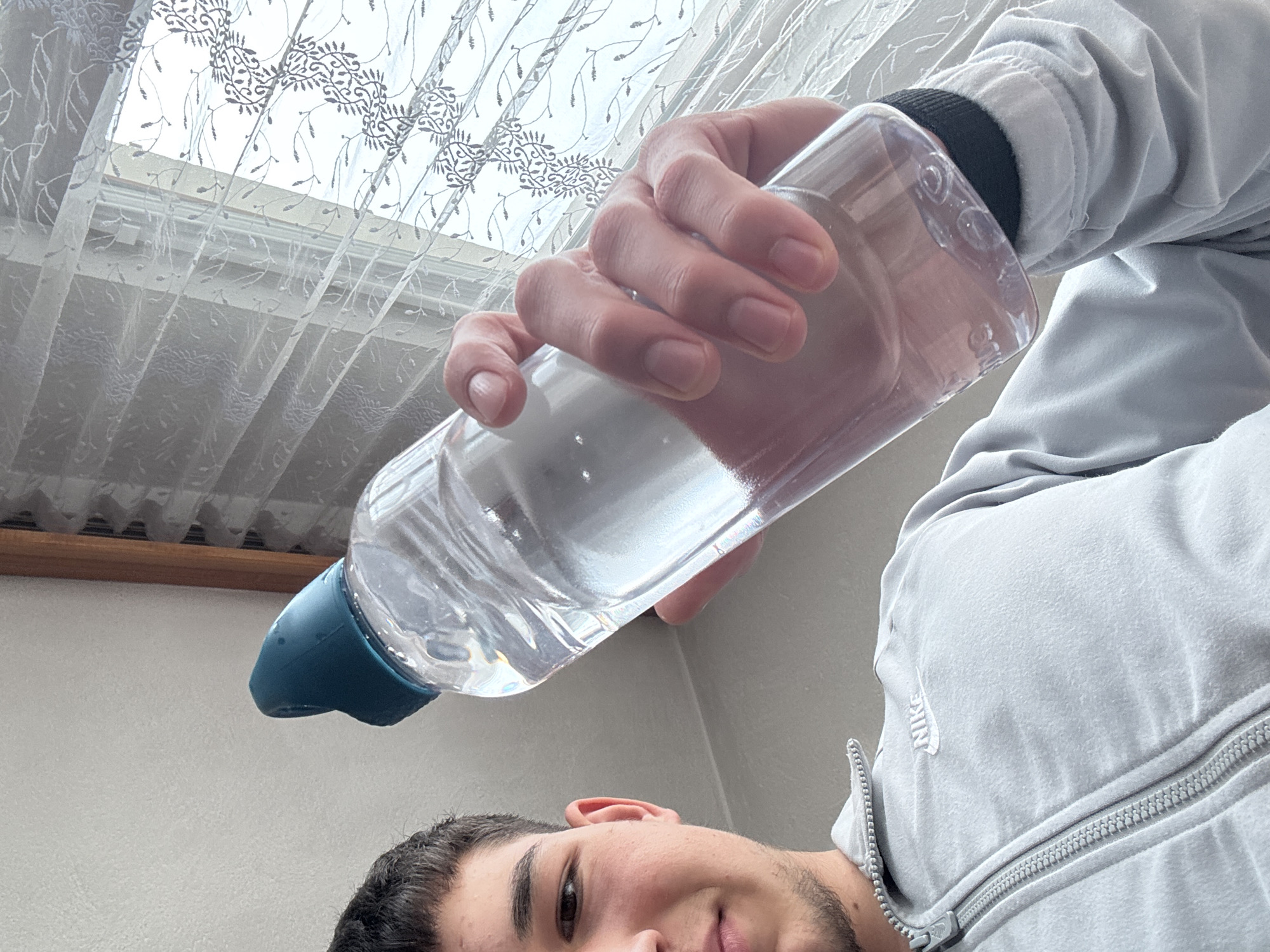 The image, captured from a low-angle perspective in Zlatograd, Bulgaria, features a young man partially visible in the lower part of the frame, looking directly upwards at the camera. He holds a clear plastic water bottle, roughly half-filled with water, in his right hand, positioned above his head. The bottle has a dark teal or blue cap and spout.
The man has dark hair and a light beard, with a neutral to slightly smiling expression. He is wearing a light grey jacket with a prominent zipper. A white embroidered "NIKE" logo is visible on the left side of his jacket.
The background reveals an indoor setting, likely a room with a window. The window is covered by a sheer white curtain adorned with intricate grey floral or leafy patterns, allowing soft, diffused daylight to enter. Parts of the window frame and a wooden sill are visible behind the translucent curtain. The bright illumination suggests it is daytime. Faint molded text, including "ml" and possibly "600", can be seen on the clear plastic bottle, indicating its volume.