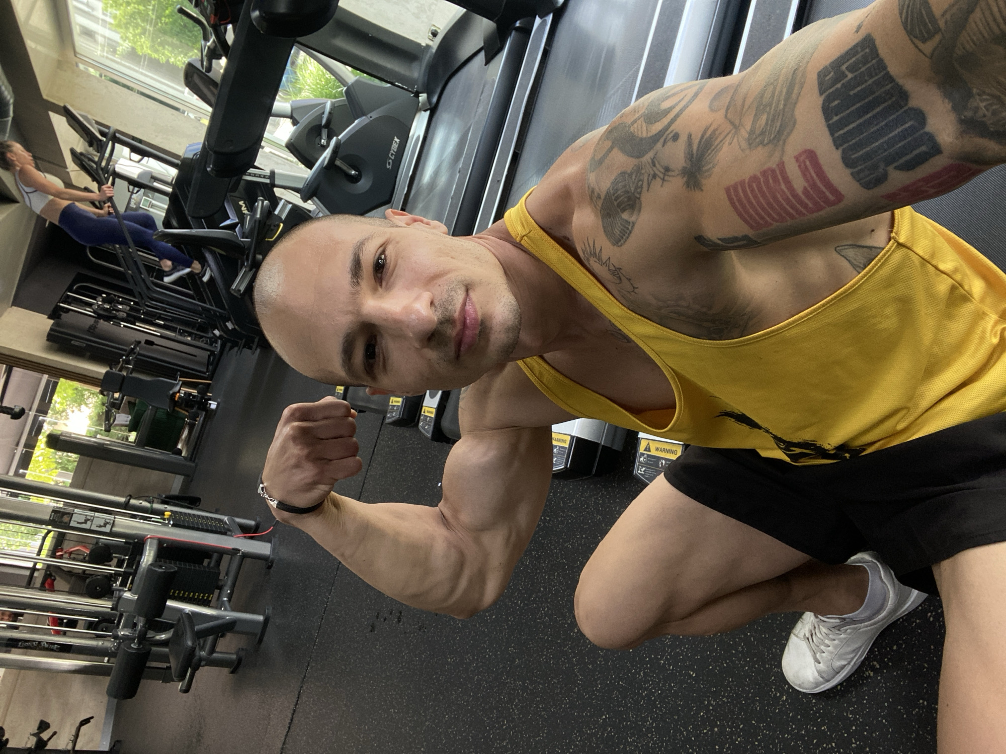This is a selfie taken by a man in a gym. He is wearing a yellow tank top and black shorts, and has his left arm flexed, showing off his bicep. He has tattoos on his right arm. In the background, other gym equipment and a woman on a treadmill are visible. The gym appears to be modern and well-equipped, with large windows looking out onto a city. The location is the XYZ Fitness Club in Pleven, Bulgaria.