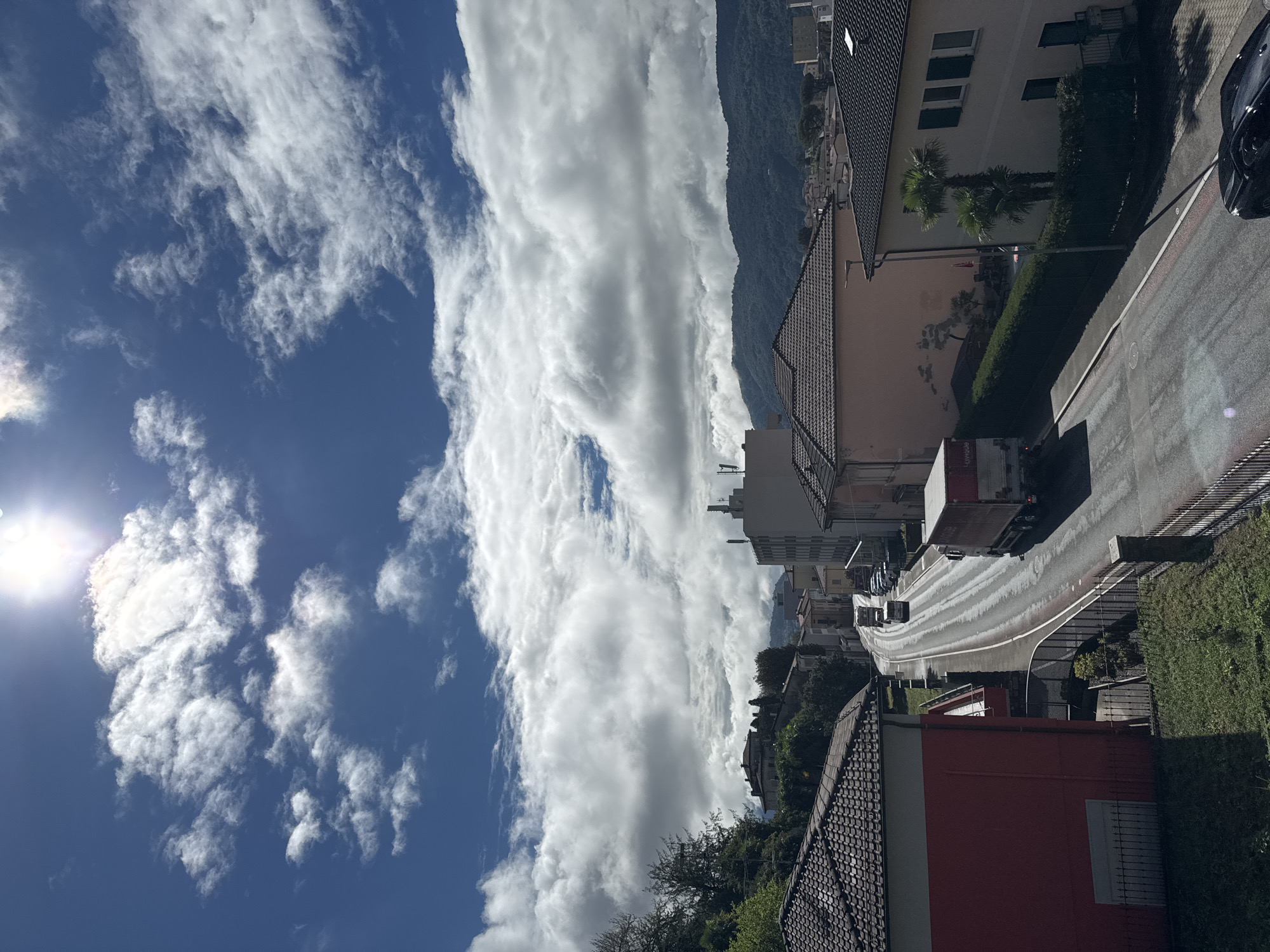 The image captures a sun-drenched street scene in Balerna, Switzerland, under a dynamic sky filled with dramatic white and gray clouds. The sun is positioned high and bright in the upper right quadrant of the sky, casting a strong glare. A two-lane road dominates the center of the frame, curving gently to the left and leading uphill.
On the road, a red and white semi-trailer truck is moving uphill, followed by a white cargo van. Several parked cars are visible along the right side of the road. To the left of the road, a vibrant red building with a dark tiled roof is prominently featured in the foreground. Further up the street, on the right side, a light-colored building with multiple windows and a dark tiled roof is visible, alongside a neatly trimmed green hedge and two palm trees.
In the background, residential buildings with tiled roofs are nestled among green trees. Beyond the buildings, rolling hills covered in dense green foliage rise towards the horizon, partially obscured by the clouds. The overall impression is a lively urban landscape bathed in bright sunlight, with the natural beauty of the surrounding hills providing a scenic backdrop.