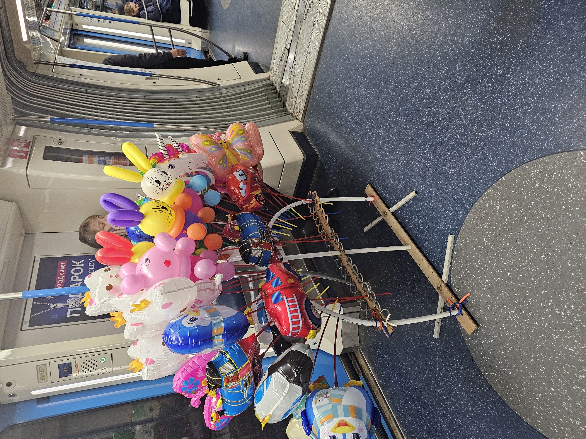 The image shows a colorful collection of balloons on a wooden rack inside what appears to be a subway car in Moscow, Russia. The balloons are shaped like various animals and characters, including rabbits, unicorns, butterflies, and cartoon cars. A few passengers are visible in the background. The interior of the subway car includes metal walls with horizontal ridges and blue speckled flooring. A poster with Cyrillic text is visible near the balloons.
